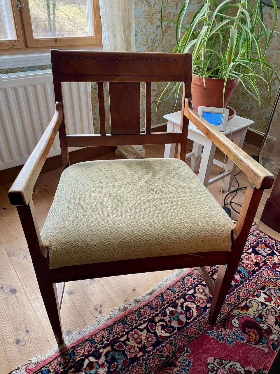 A wooden armchair with beige patterned upholstery on the seat, placed on a colorful Persian-style rug in a room with wood flooring. Behind the chair, there is a window, a white radiator, a potted plant on a small white table, and a digital clock.