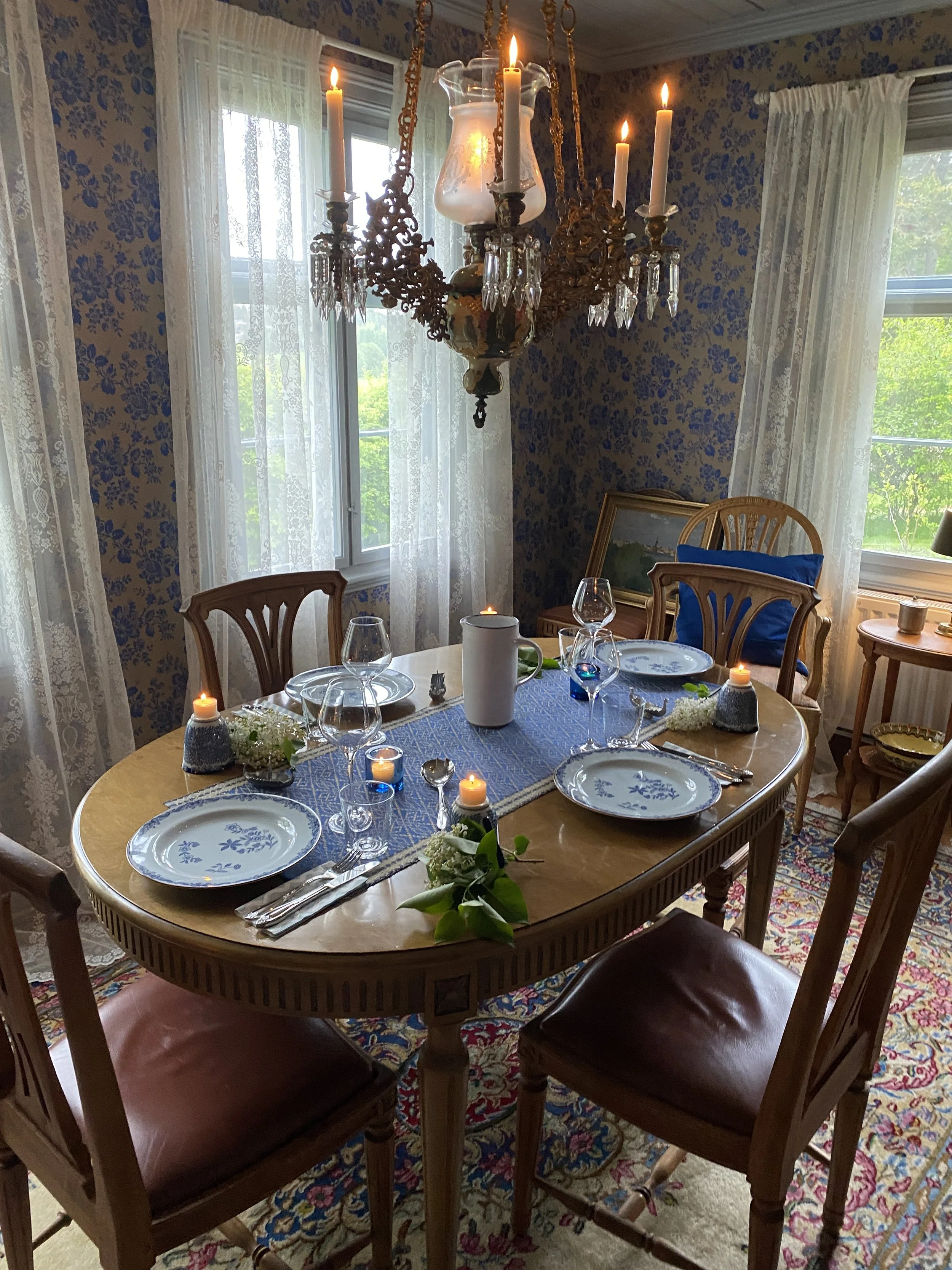 A cozy dining room set for a meal with a wooden oval table, four chairs, and a chandelier with lit candles hanging above. The table has plates, wine glasses, silverware, candles, flowers, and a blue table runner. There are windows with lace curtains 