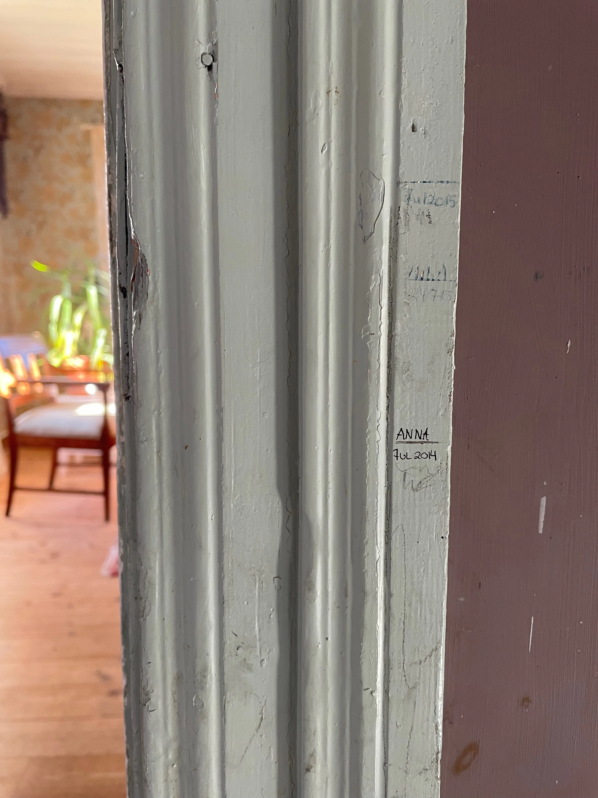 Close-up of a painted door frame with handwritten notes and date on it, with a blurry background showing a wooden chair and potted plant.