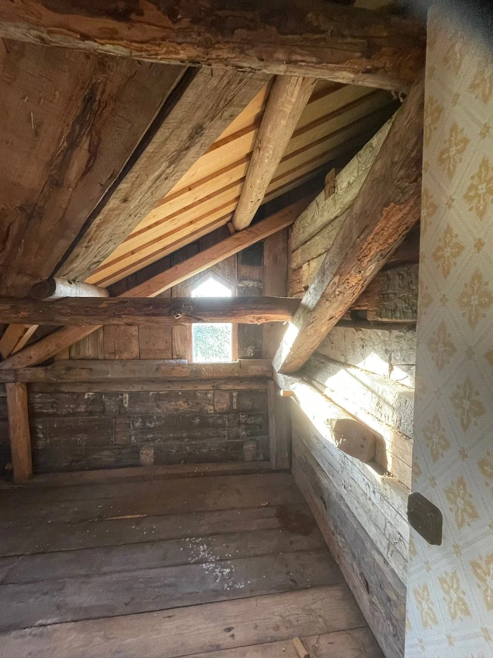 Interior view of an attic or loft space showing wooden beams, old weathered walls, and new wooden roof trusses with sunlight coming through small windows.
