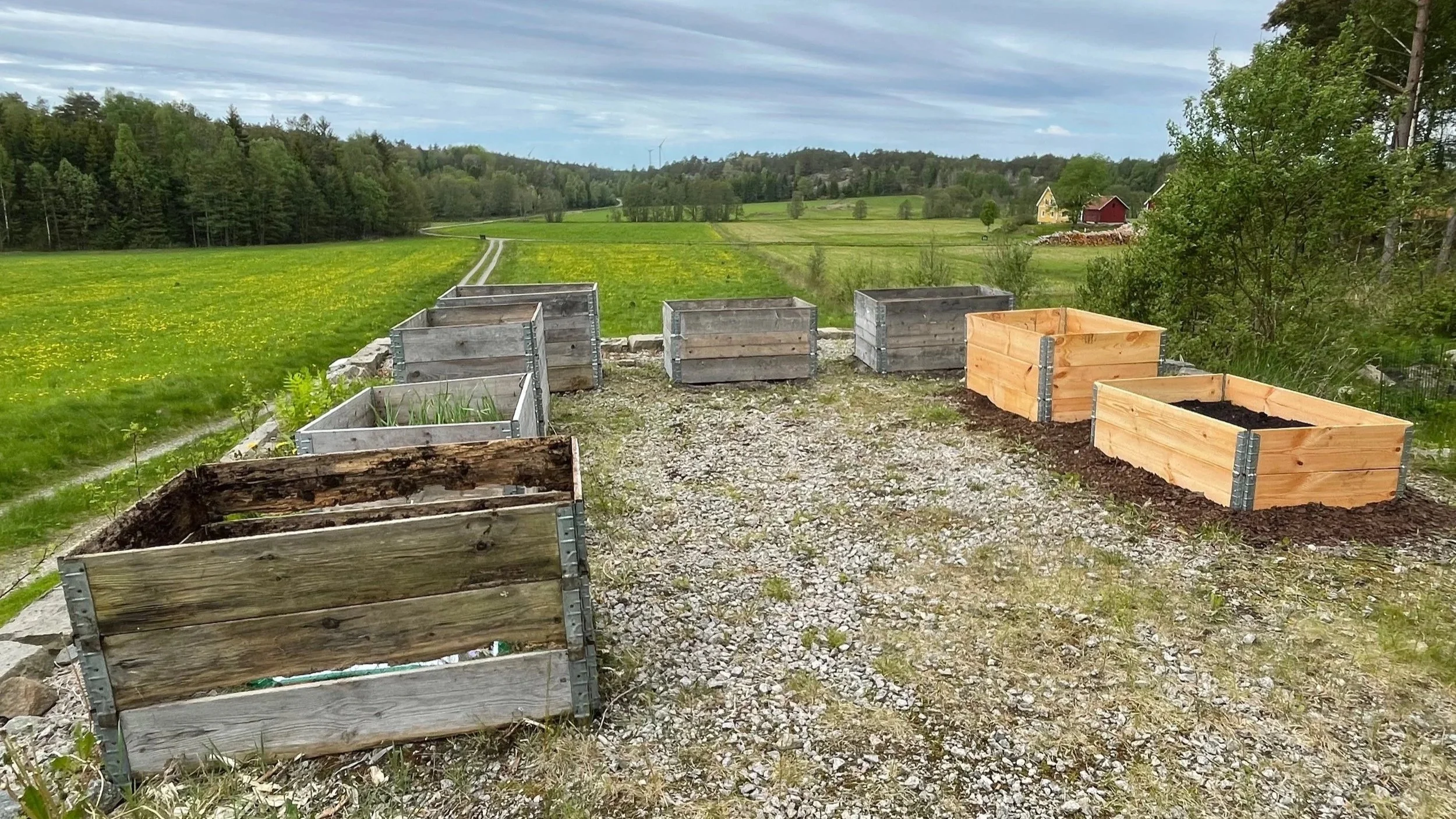 View of a countryside landscape with several empty wooden garden planters on a gravel path, green fields, trees, houses, and cloudy sky in the background.