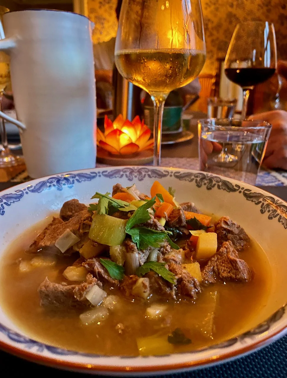 A bowl of beef stew with vegetables, garnished with fresh herbs, on a dining table set with wine and water glasses, a lit candle, and a pitcher in the background, in a cozy restaurant setting.