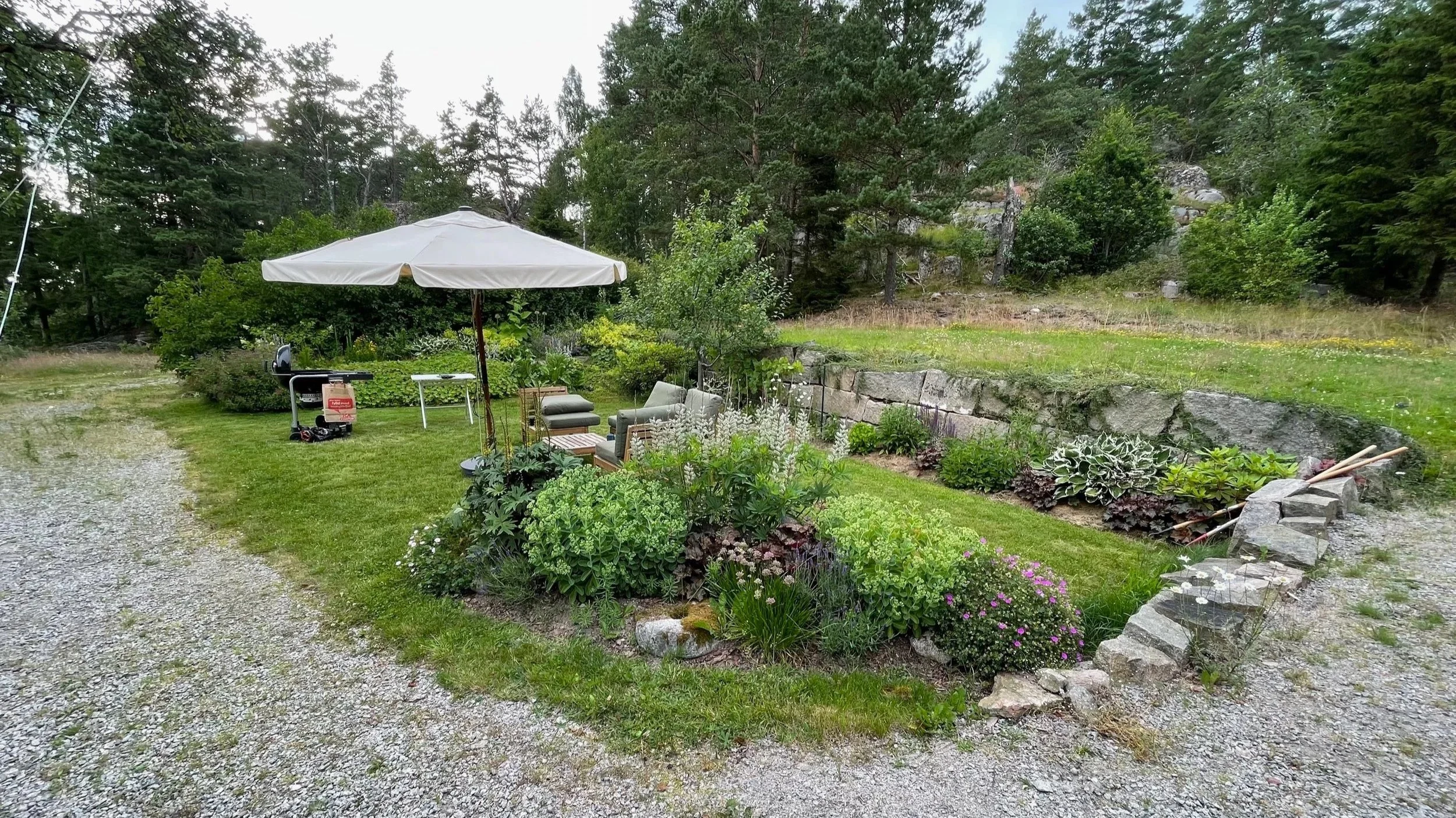 An outdoor garden area with patio furniture, including a table and chairs, under a white umbrella. There are various plants and flowers along a stone border, with a wooded hillside in the background.