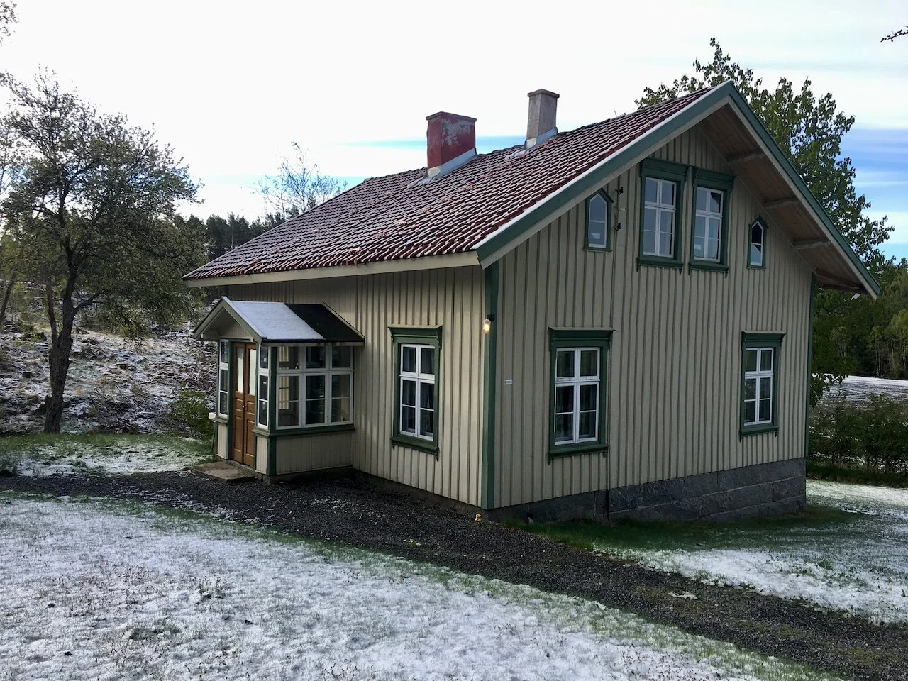 A two-story house with a red tiled roof, yellow vertical wood siding, and green window trims, situated on a small hill with some snow on the ground and trees around.