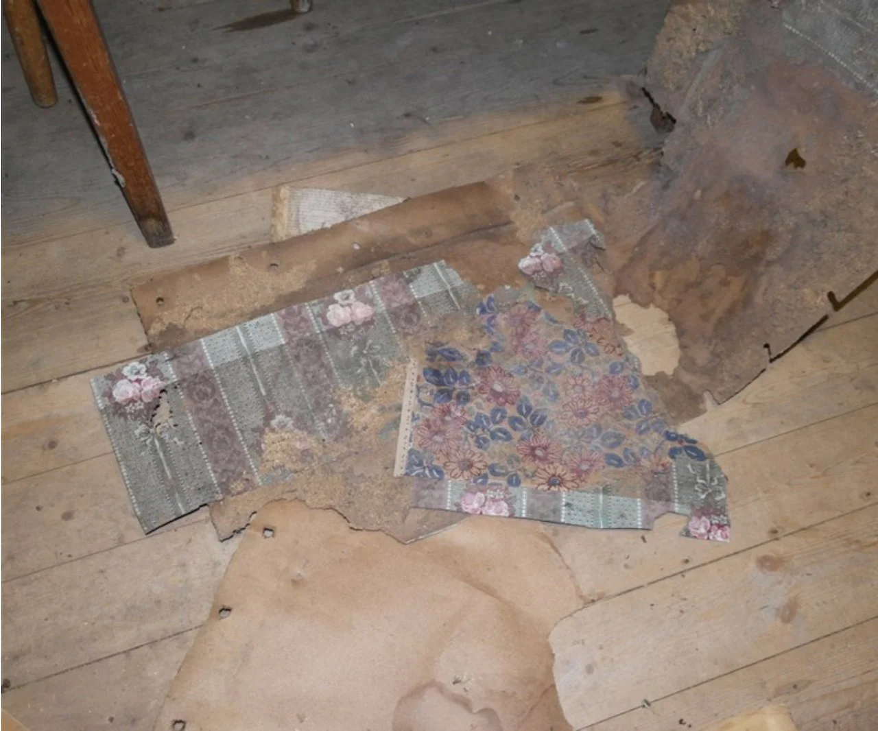 Damaged and torn floral wallpaper on wooden floor with exposed subfloor underneath.