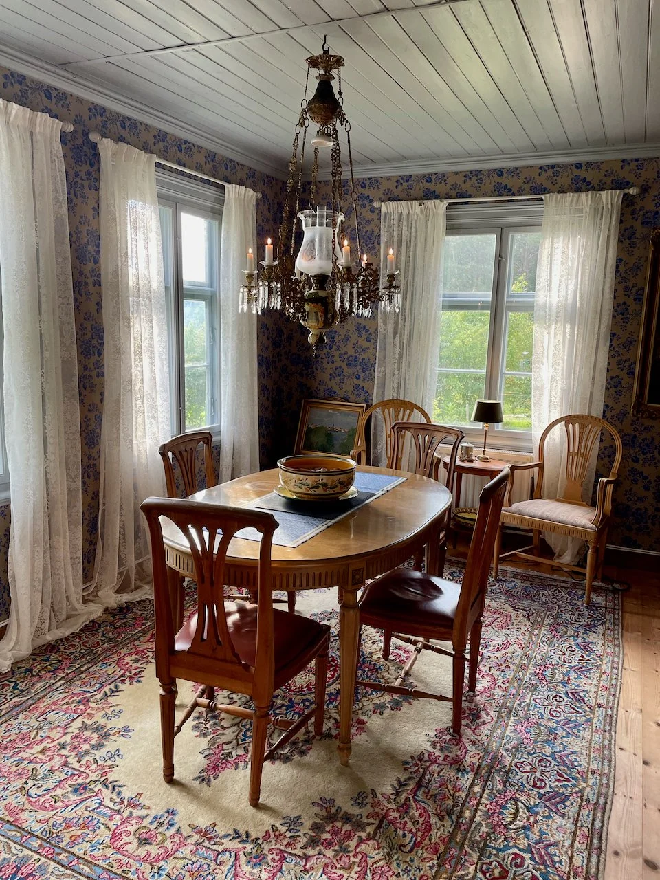 A vintage dining room with a wooden oval table, six chairs, lace curtains, and floral wallpaper. A crystal chandelier hangs above, and sunlight streams through two windows. The table has a large ceramic bowl and a placemat, and there is a small lamp and framed landscape on a side table near the window.
