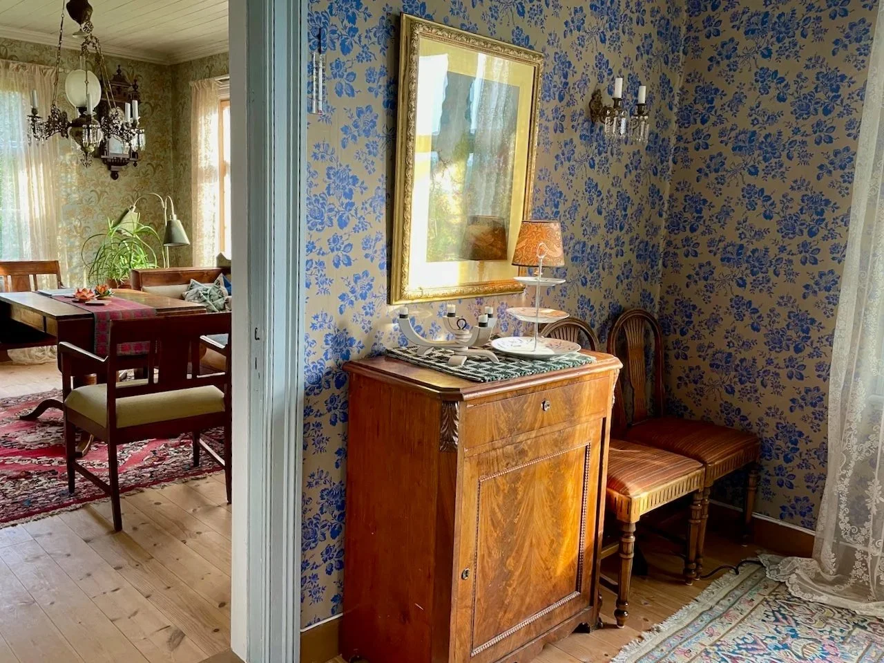 A vintage-style dining room with floral wallpaper, a wooden sideboard, two chairs, a candelabra, a table lamp with a patterned shade, and lace curtains.