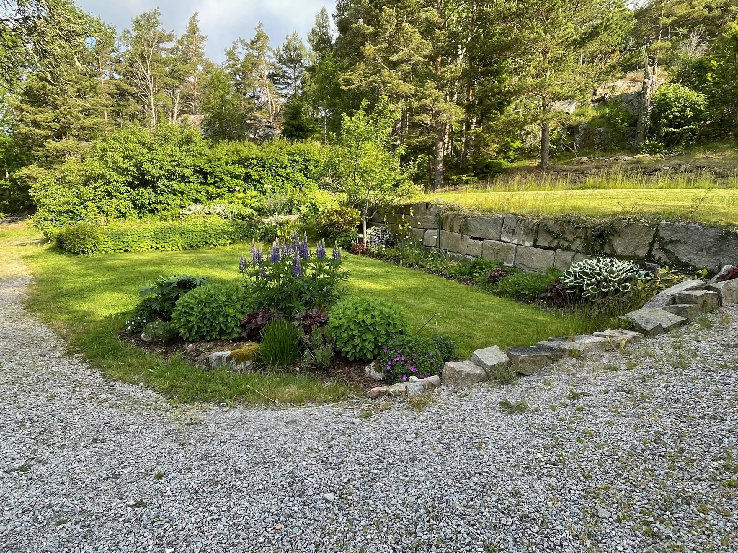 A well-maintained garden with a gravel pathway, green grass, colorful flowers, and trees in the background under a partly cloudy sky.