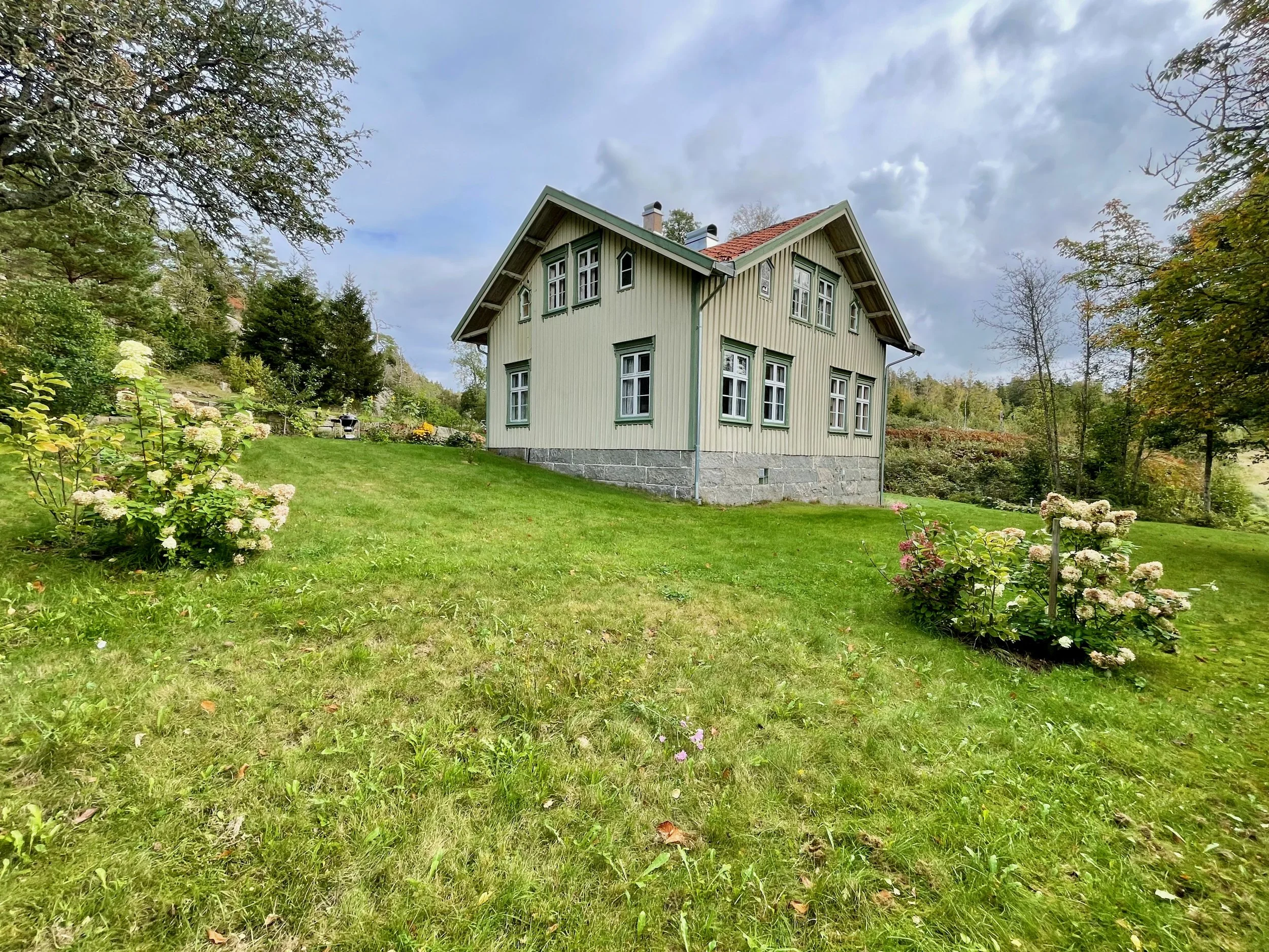 A light green multi-story house with a red tile roof, surrounded by green trees and shrubs on a slightly overcast day.