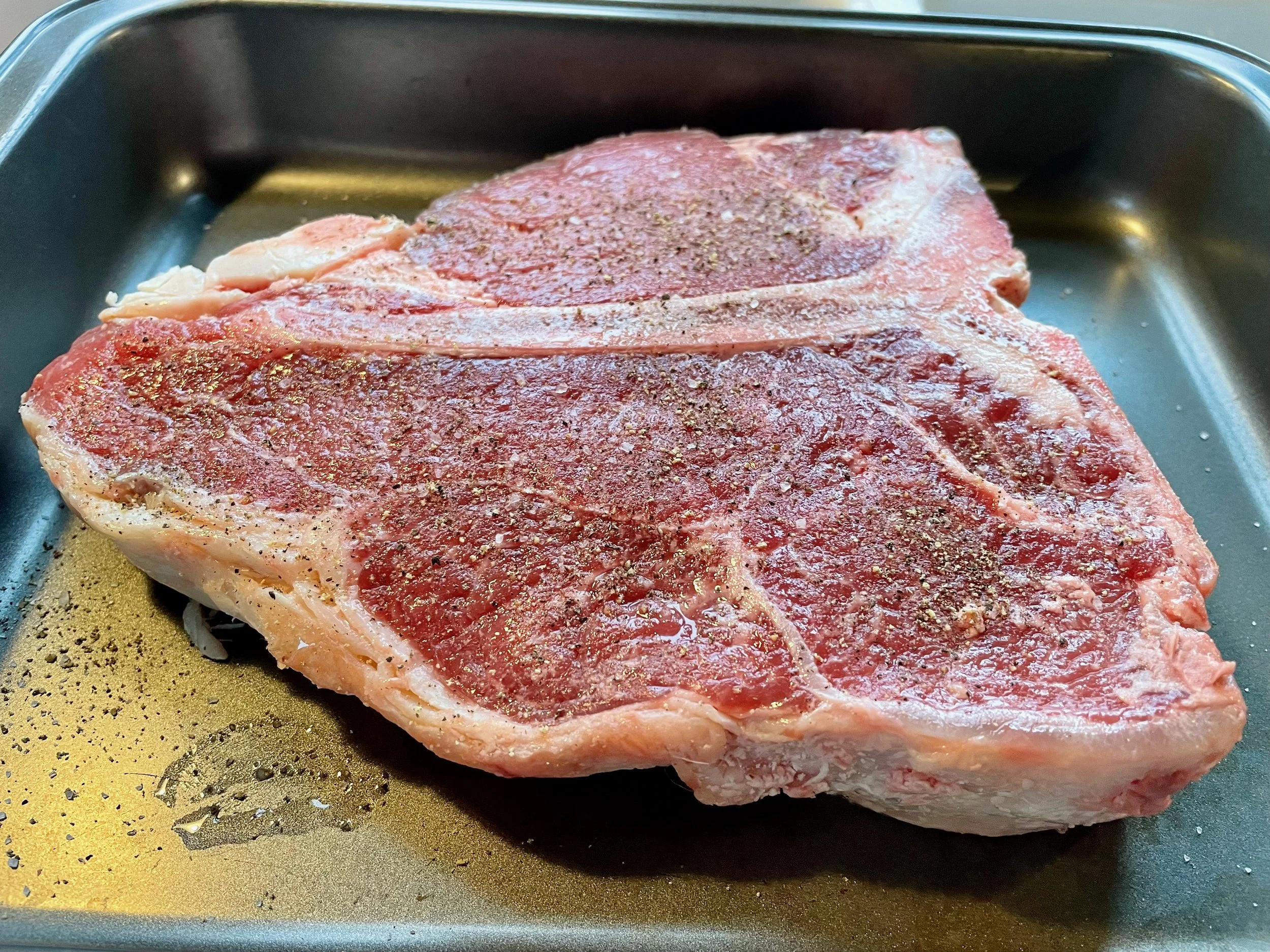 Uncooked, seasoned T-bone steak on a black baking sheet.