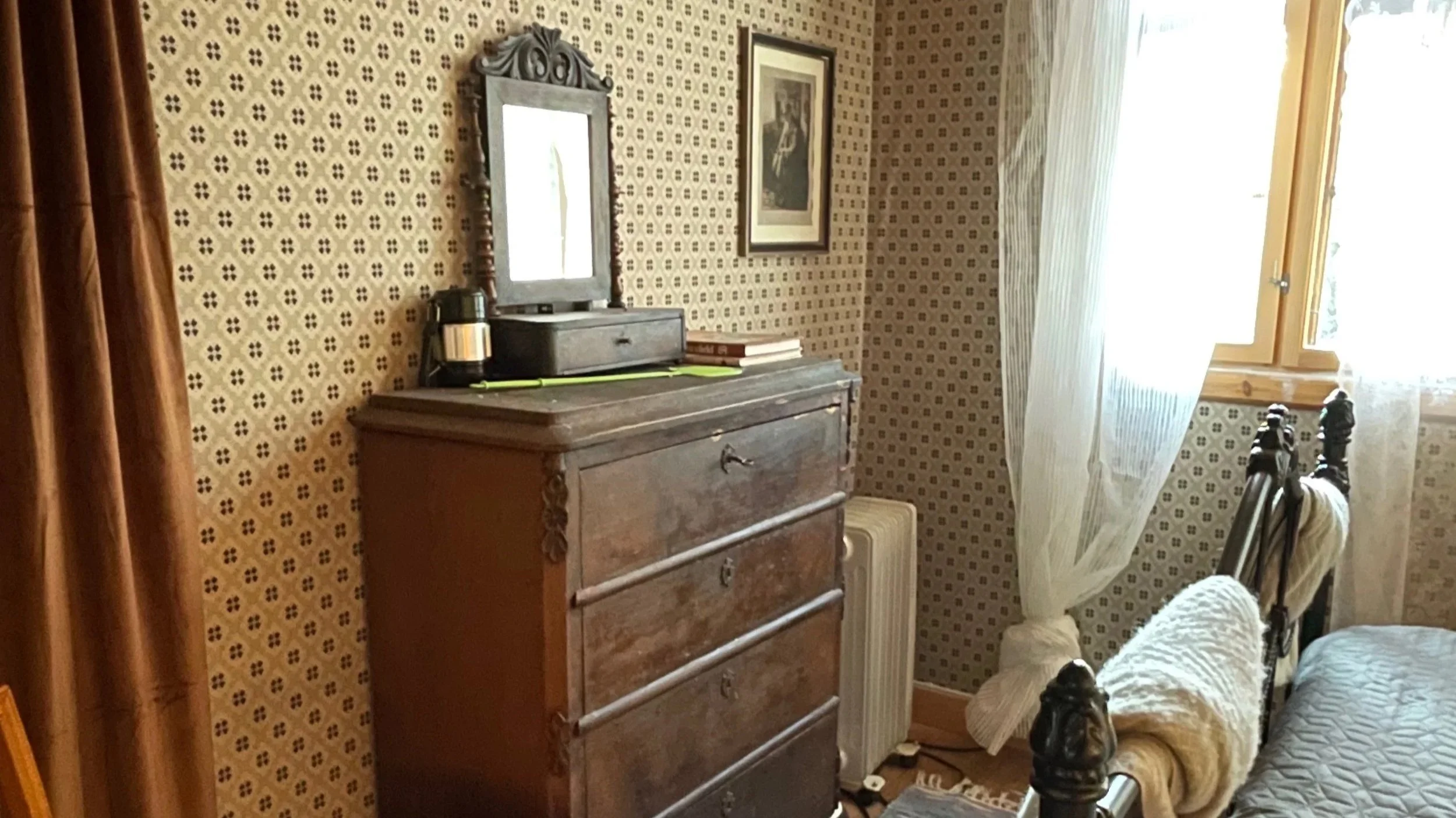 A cozy bedroom corner with a wooden dresser, a mirror, a framed picture on the wall, a window with curtains, and a bed with a rolled blanket.