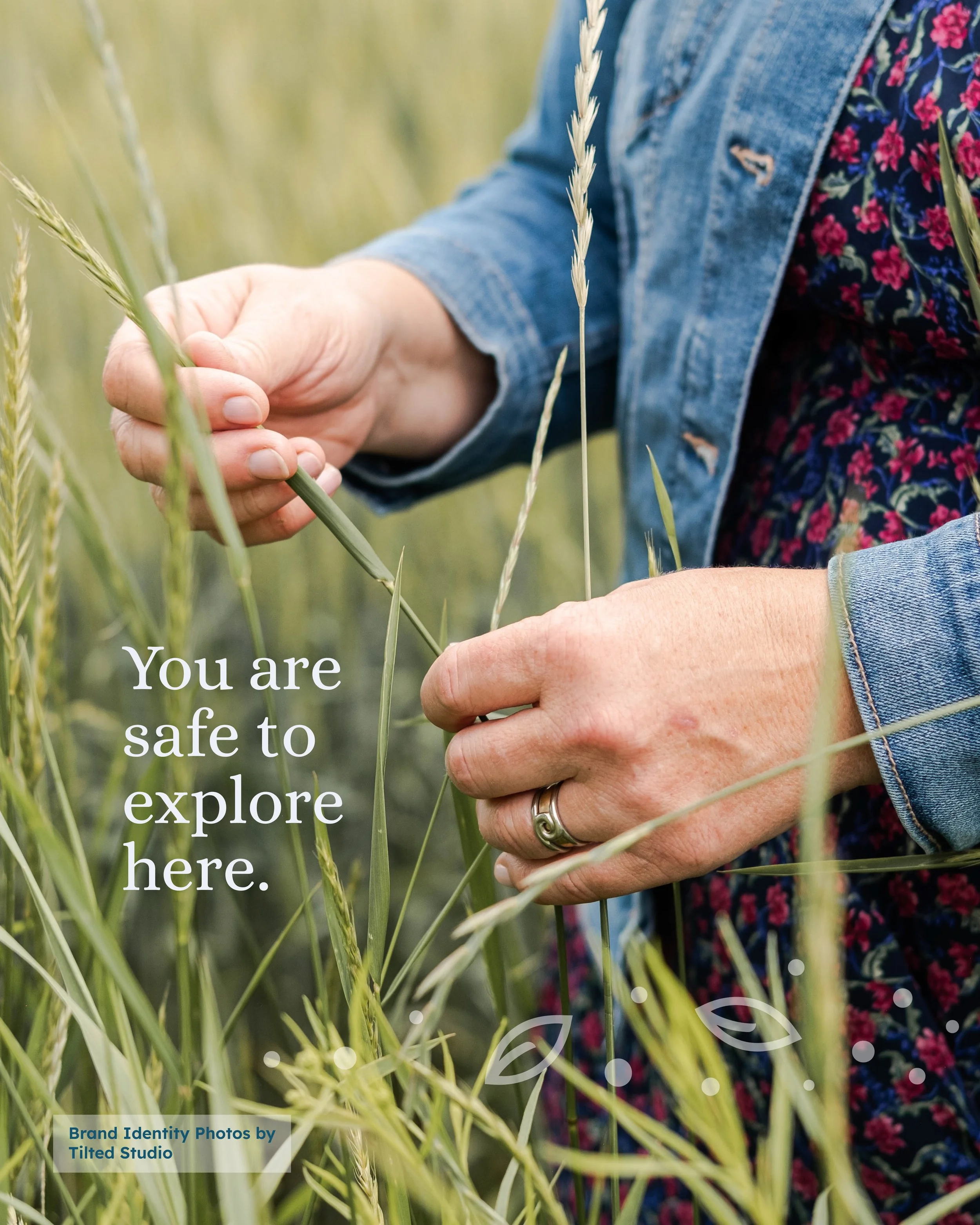 Close-up of a person in a denim jacket and floral dress exploring a grassy field, holding and closely inspecting a stalk of grass. Overlaid text reads 'You are safe to explore here'.