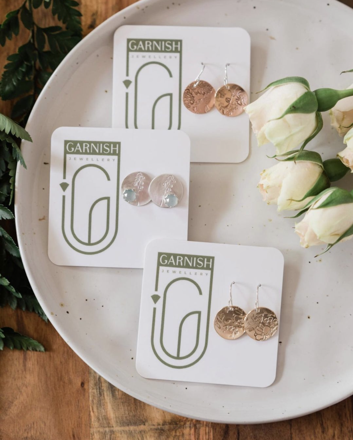 Three pairs of earrings displayed on Garnish Jewellery cards, placed on a white ceramic plate with white roses and green foliage around it.