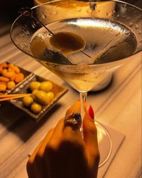 A person holding a martini glass with a green olive on a toothpick, garnished with a cherry, at a restaurant or bar setting. In the background, there is a plate with green olives and some yellow snacks.