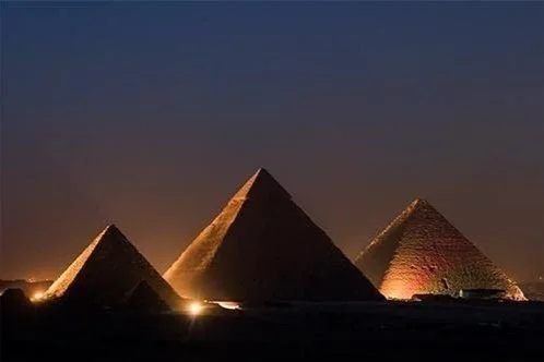 Nighttime scene of the three pyramids of Giza illuminated, with a dark sky in the background.