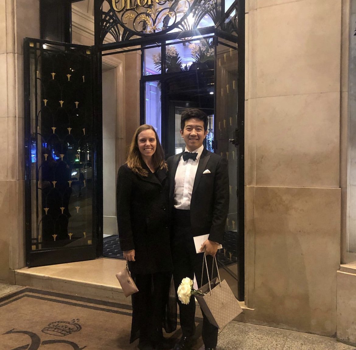 A man and woman dressed in formal attire standing outside a hotel entrance, smiling at the camera. The man is wearing a tuxedo and holding flowers, gifts, and a small purse. The woman is wearing a black dress and holding a purse.