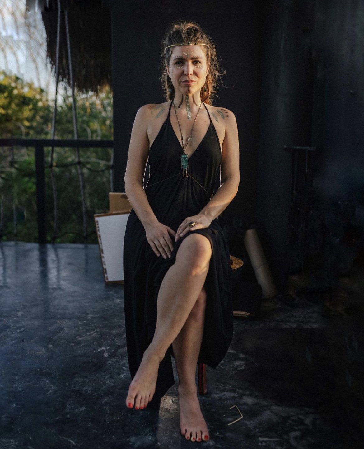 A woman sitting indoors, wearing a black dress, with tattoos, jewelry, and a headband, holding one leg up and sitting on a stool.