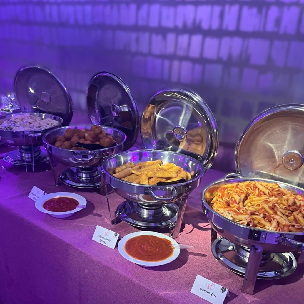 Buffet table with chafing dishes containing different types of pasta and fried food, with small bowls of marinara sauce labeled 'Mozzarella Sticks' and 'Baked Ziti' in front.