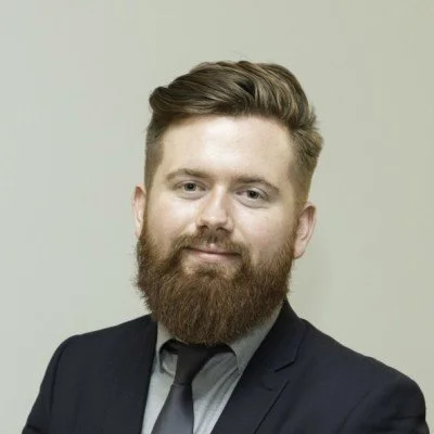 Professional headshot of a man with a beard and styled hair, wearing a dark suit and tie, against a plain background.