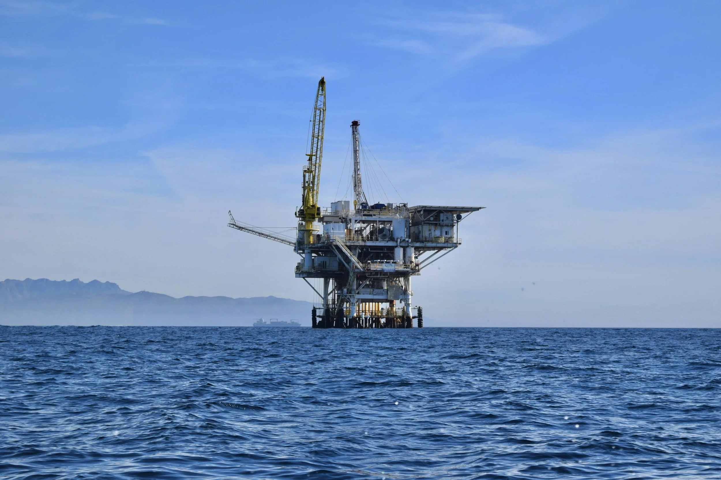 An offshore oil rig situated in the ocean with mountains in the background under a clear blue sky.