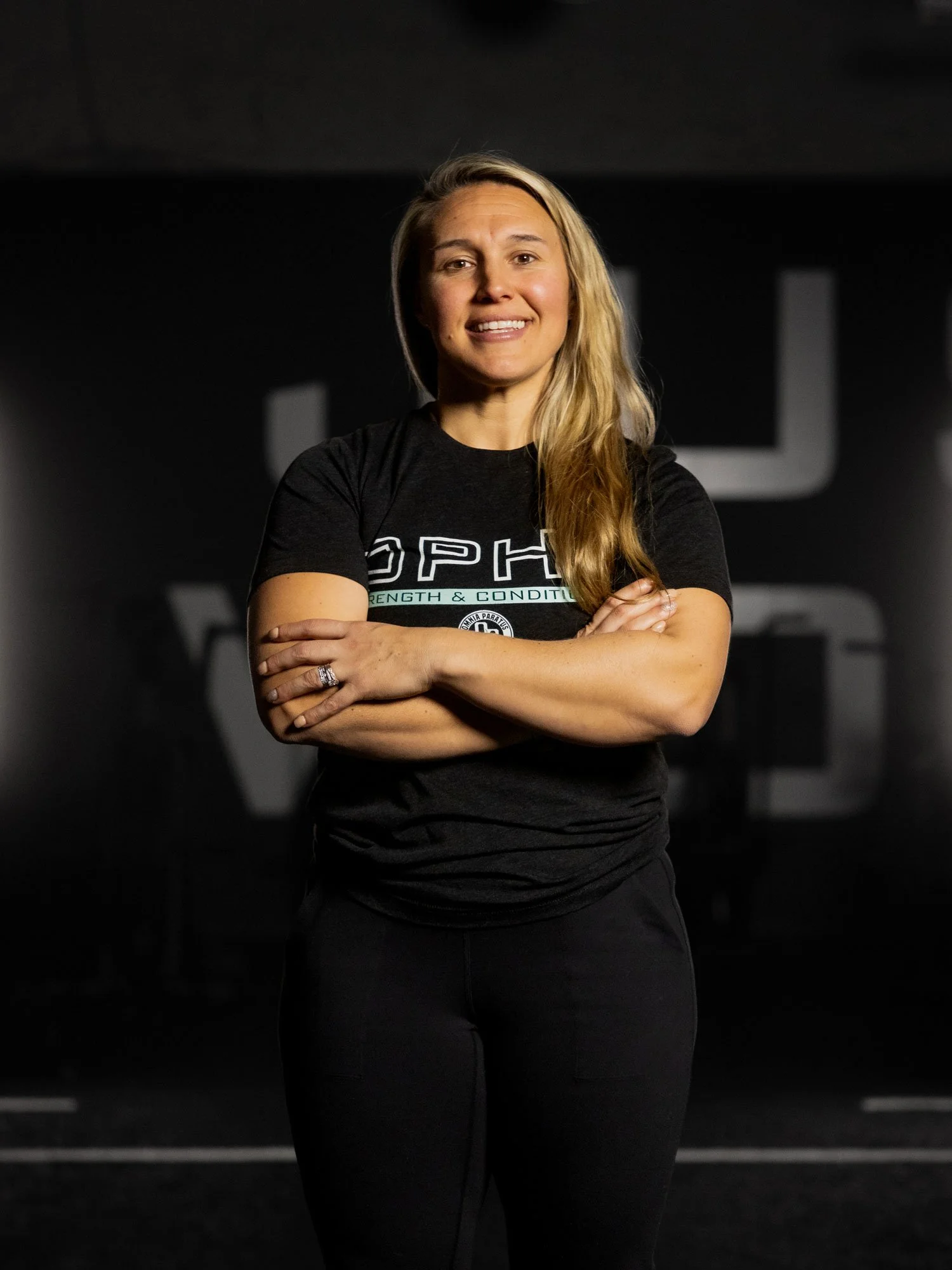 A woman with long blonde hair standing with arms crossed, smiling, wearing a black t-shirt with white and green text, in a dimly lit gym