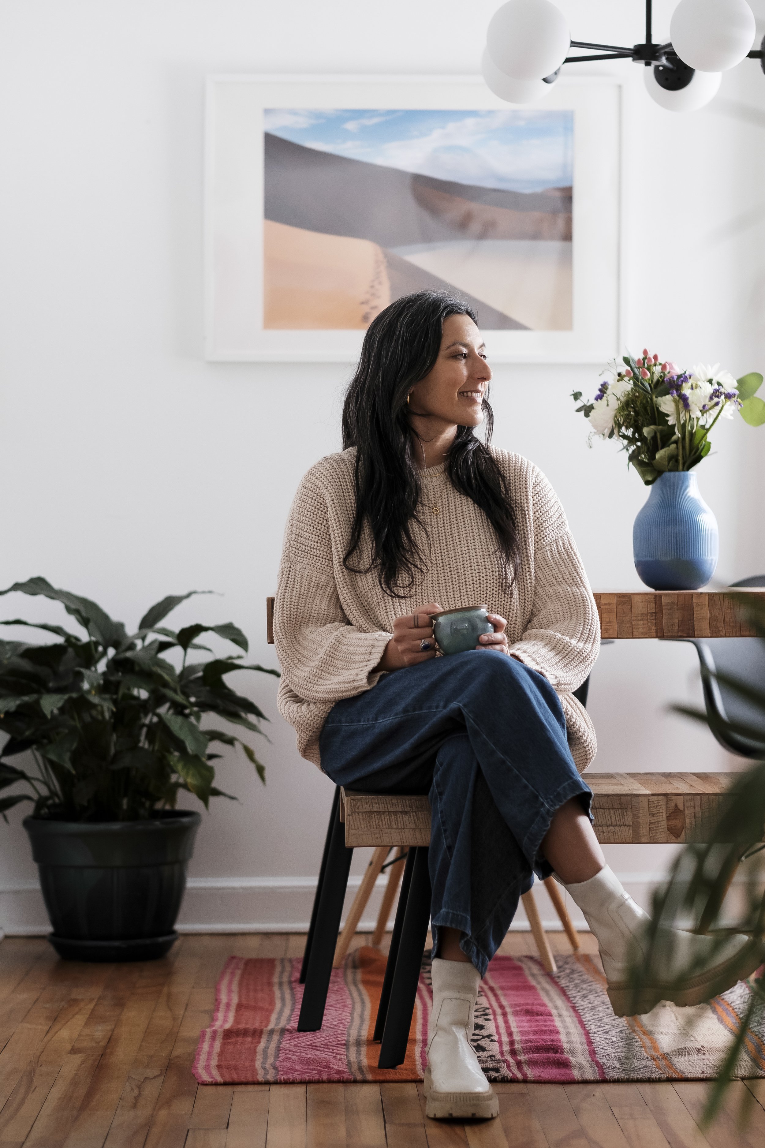 A woman with black hair, smiling, sitting on a chair in a living room, holding a mug, wearing a beige sweater, blue jeans, and white boots. There is a potted plant on the floor, a wooden table with a blue vase with flowers, a framed landscape picture on the white wall, and a modern chandelier overhead.