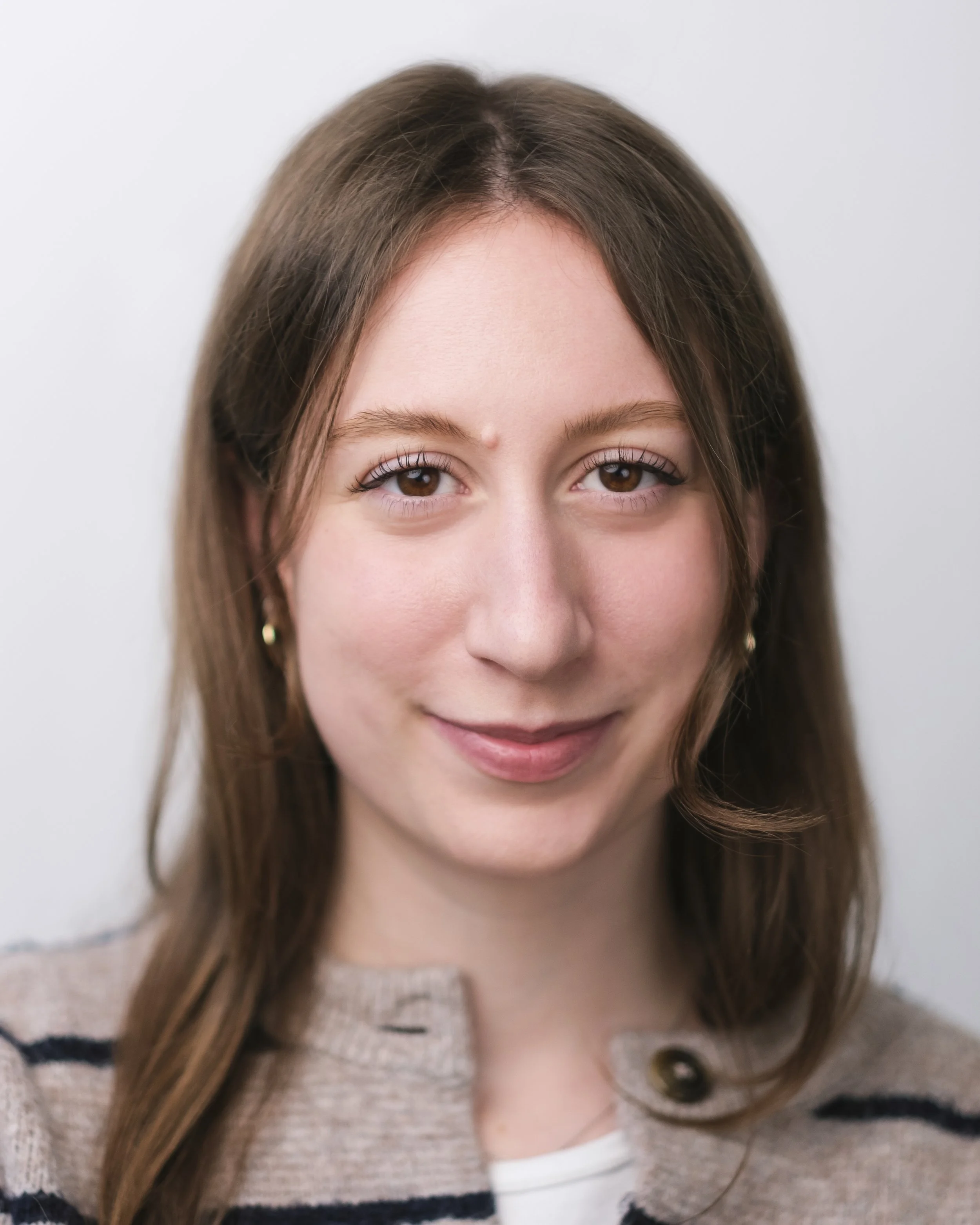 Close-up portrait of a young woman with brown hair, hazel eyes, and a light complexion, smiling gently against a plain white background.