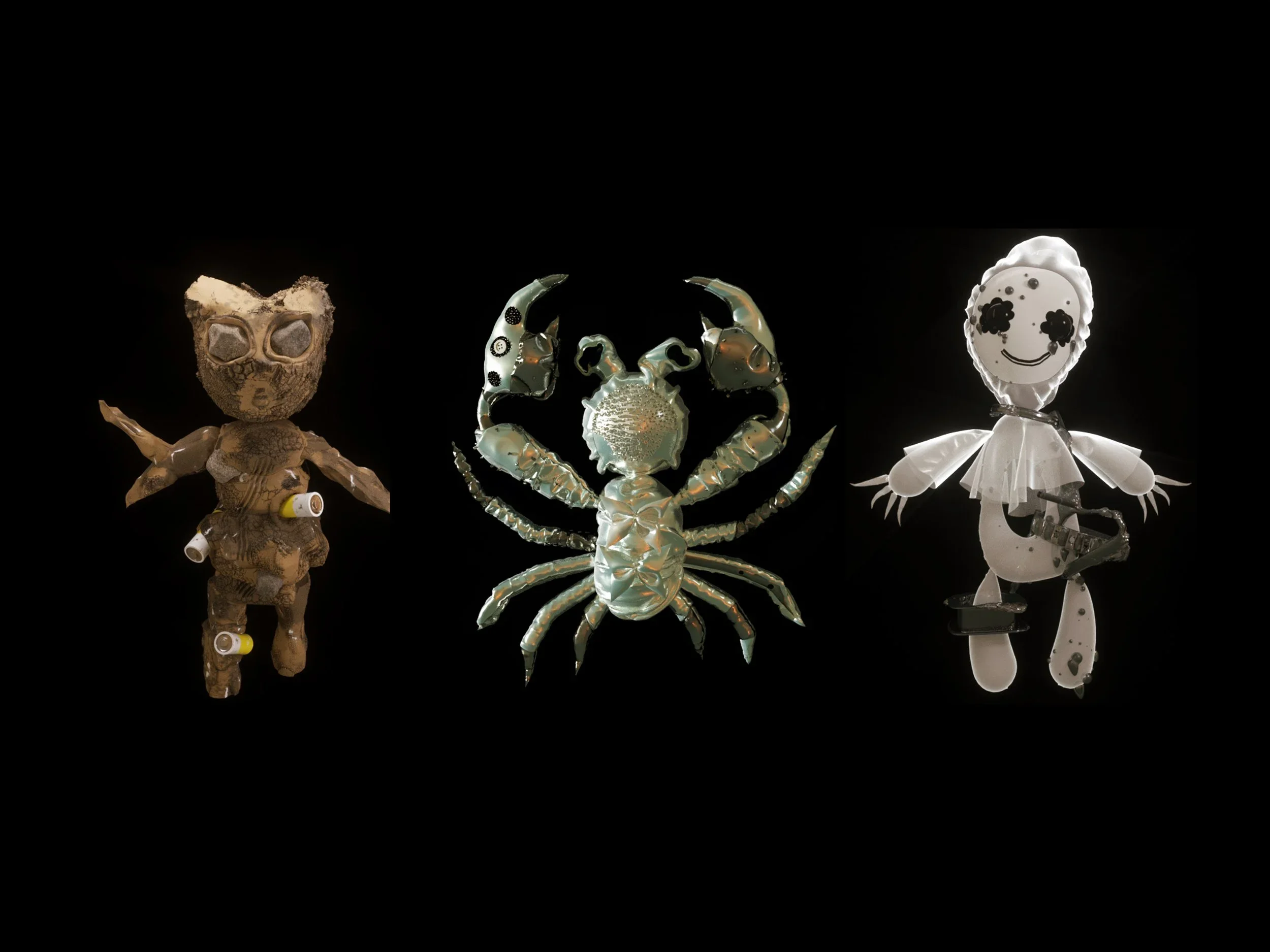 Three sculptures resembling humanoid figures made from various materials against a black background. The left figure has a foam head with glasses and a body made from textured material with small tubes. The middle figure is a metallic crab with detailed claws and legs. The right figure has a round face with a drawn smile and black eyes, wearing a tutu-like skirt, with limbs made from assorted materials.