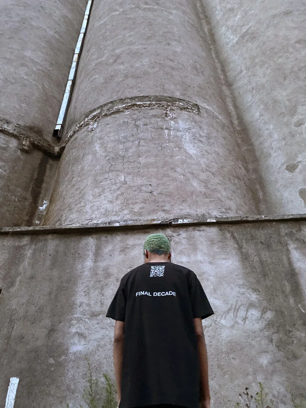 Person with green hair wearing a black t-shirt with a QR code and the words 'FINAL DECADE' printed on the back, standing with their back to the camera against a large, weathered concrete wall or structure.