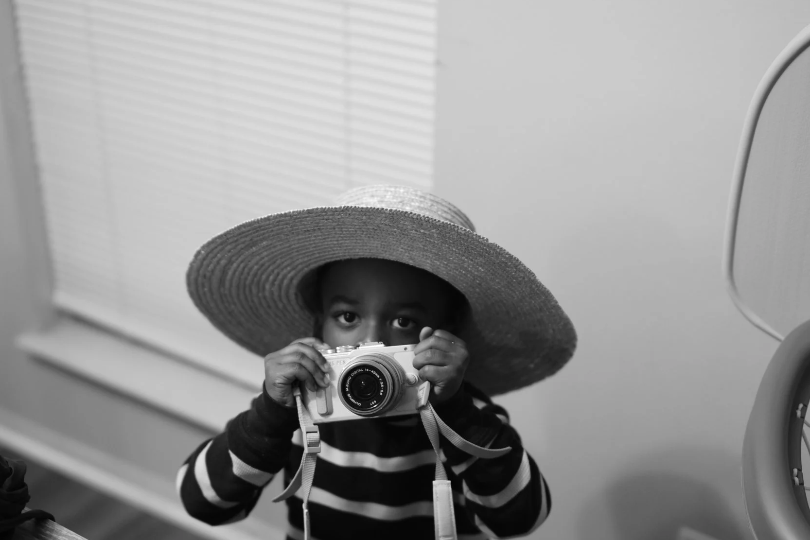 A young child wearing a large straw hat is holding a camera and looking directly at the camera.