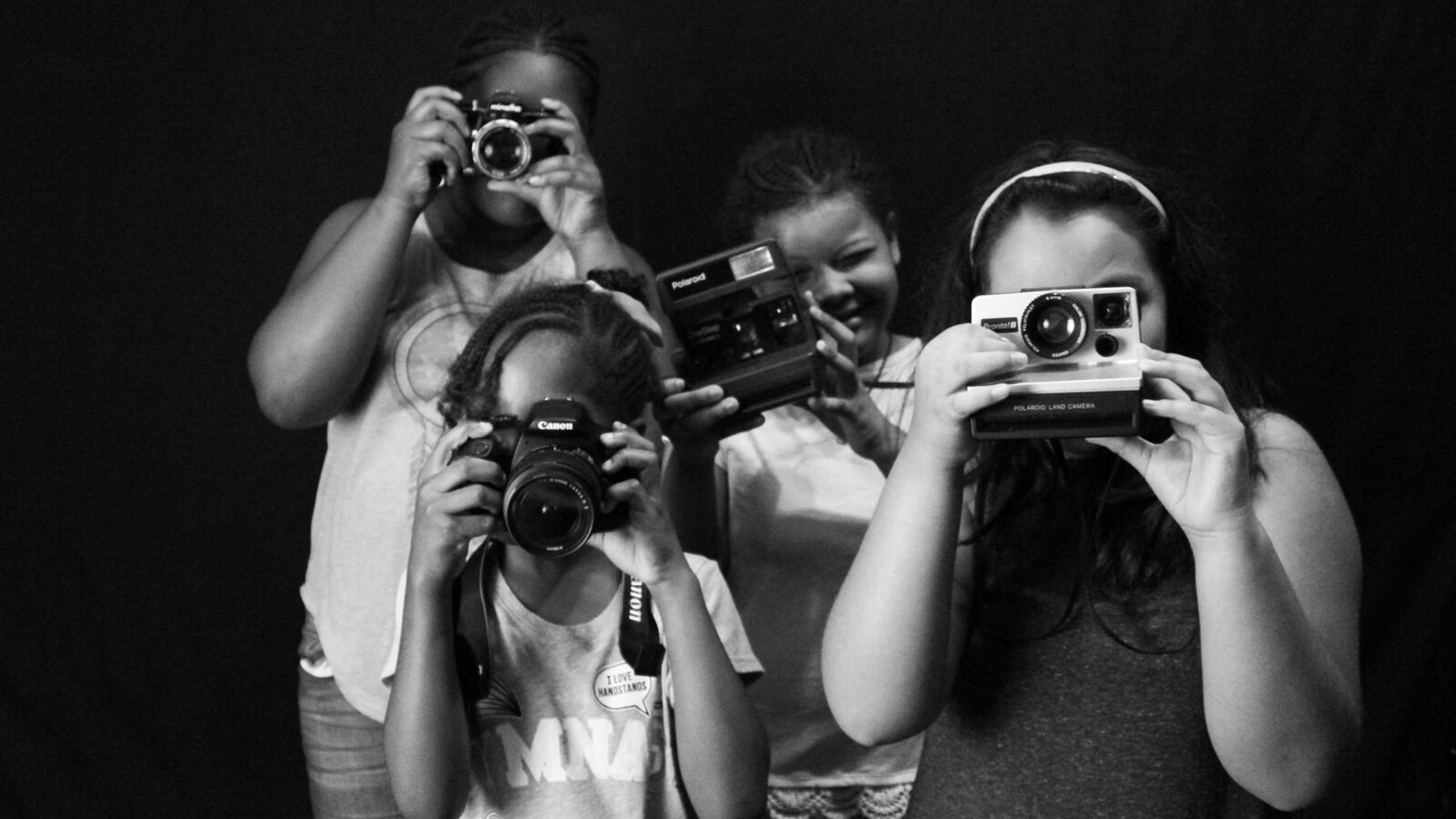 Four children holding cameras, taking pictures, with a dark background.