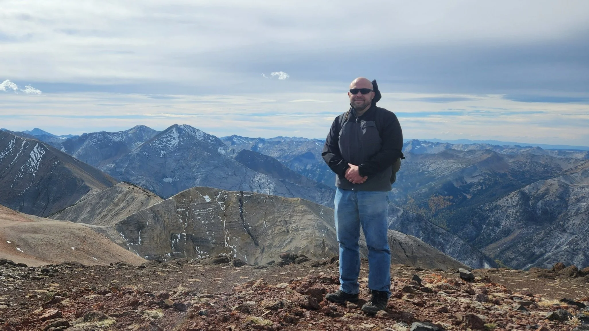 Person standing on a rocky mountain peak with snowy, rugged mountains in the background.
