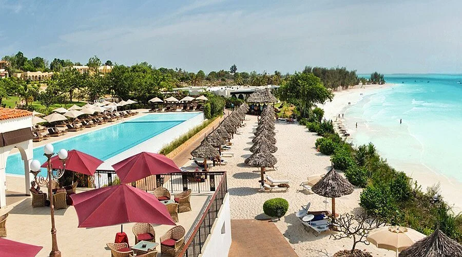A beachside resort with lounge chairs, umbrellas, a swimming pool, and a sandy beach with ocean waves.