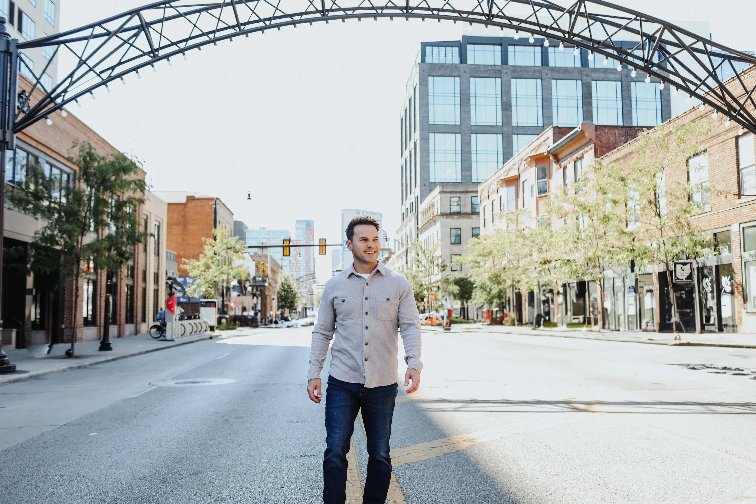 Brian walking in the middle of a city street under a metal archway, with tall buildings on either side, trees lining the sidewalks, and traffic lights in the background.