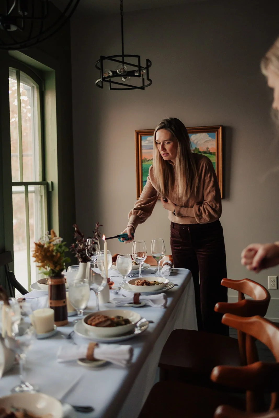 Woman lighting a candle at a dinner table with plates, wine glasses, and a floral centerpiece in a well-lit dining room.