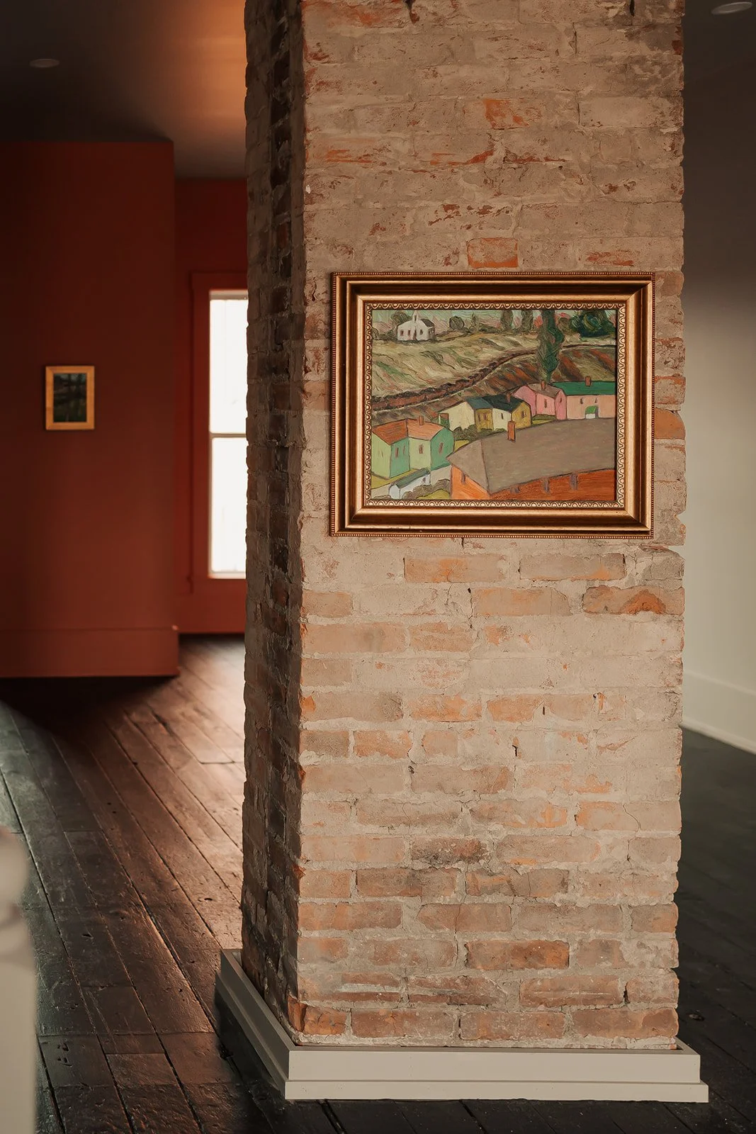 A framed painting of a colorful hillside village hangs on a brick column inside a house, with a window and a red wall visible in the background.