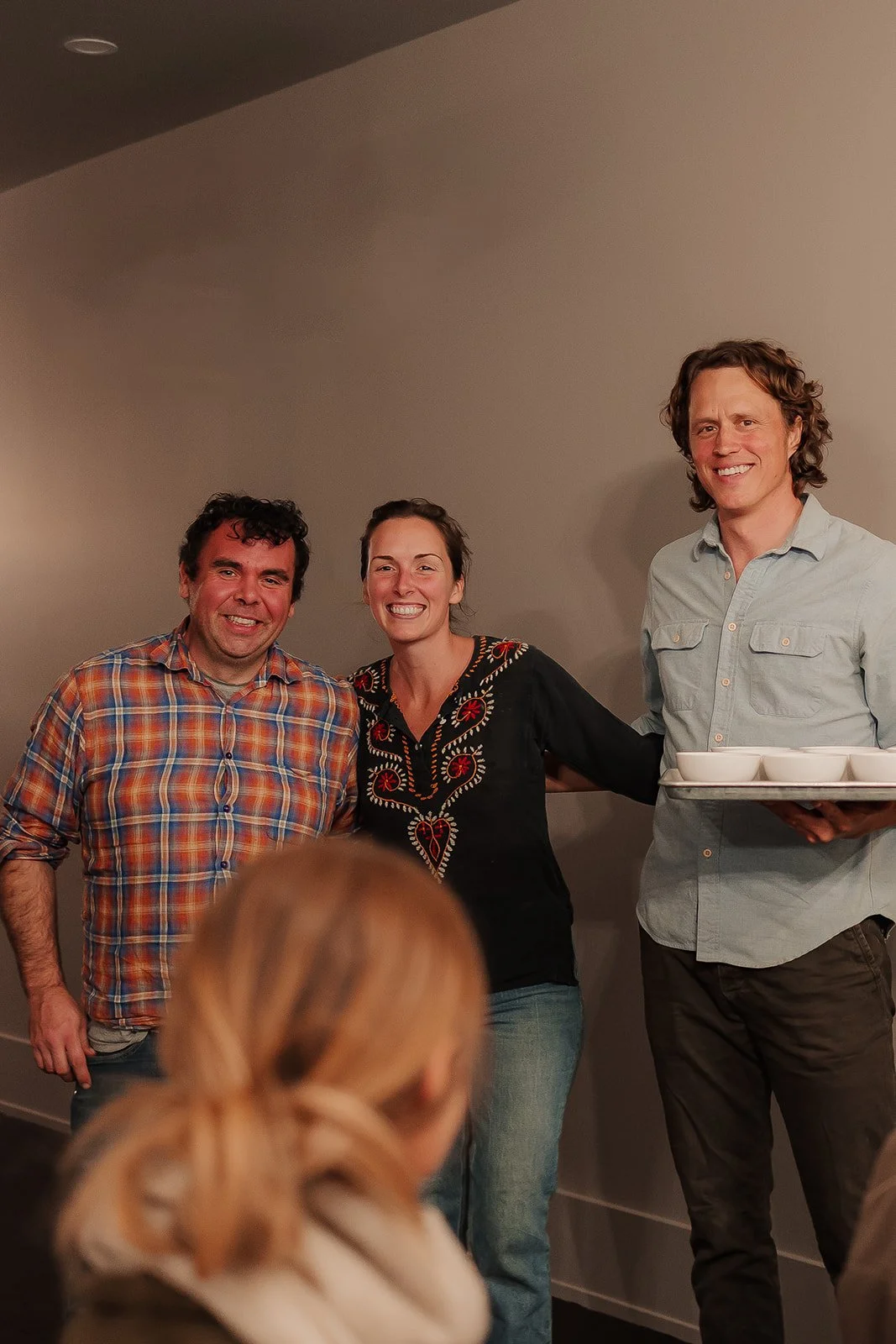 Three cheerful people stand together indoors, posing for a photo, with a woman in the foreground facing away. The man on the right holds a tray with four small bowls.