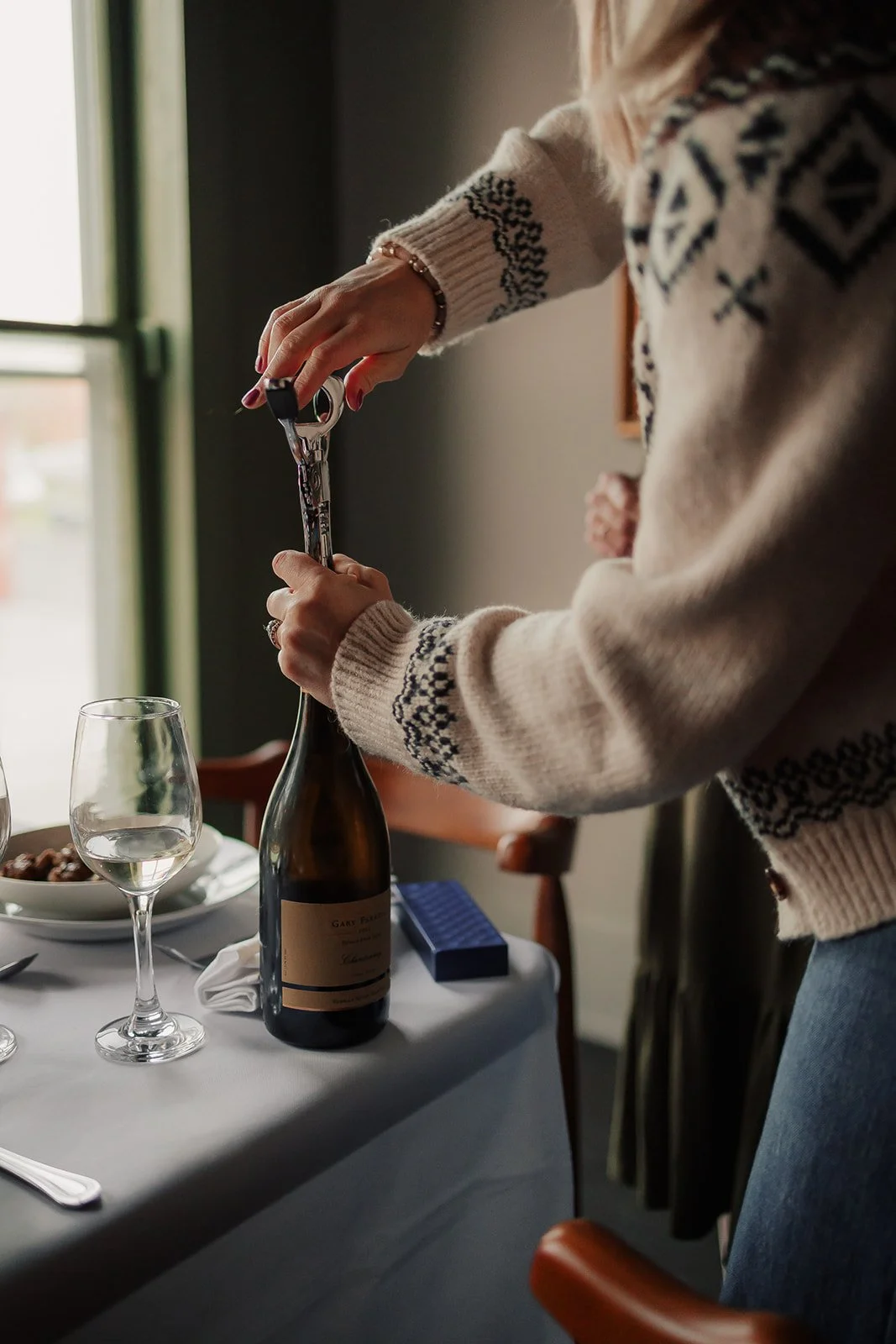 Person opening a bottle of wine at a dining table with wine glasses and plates.