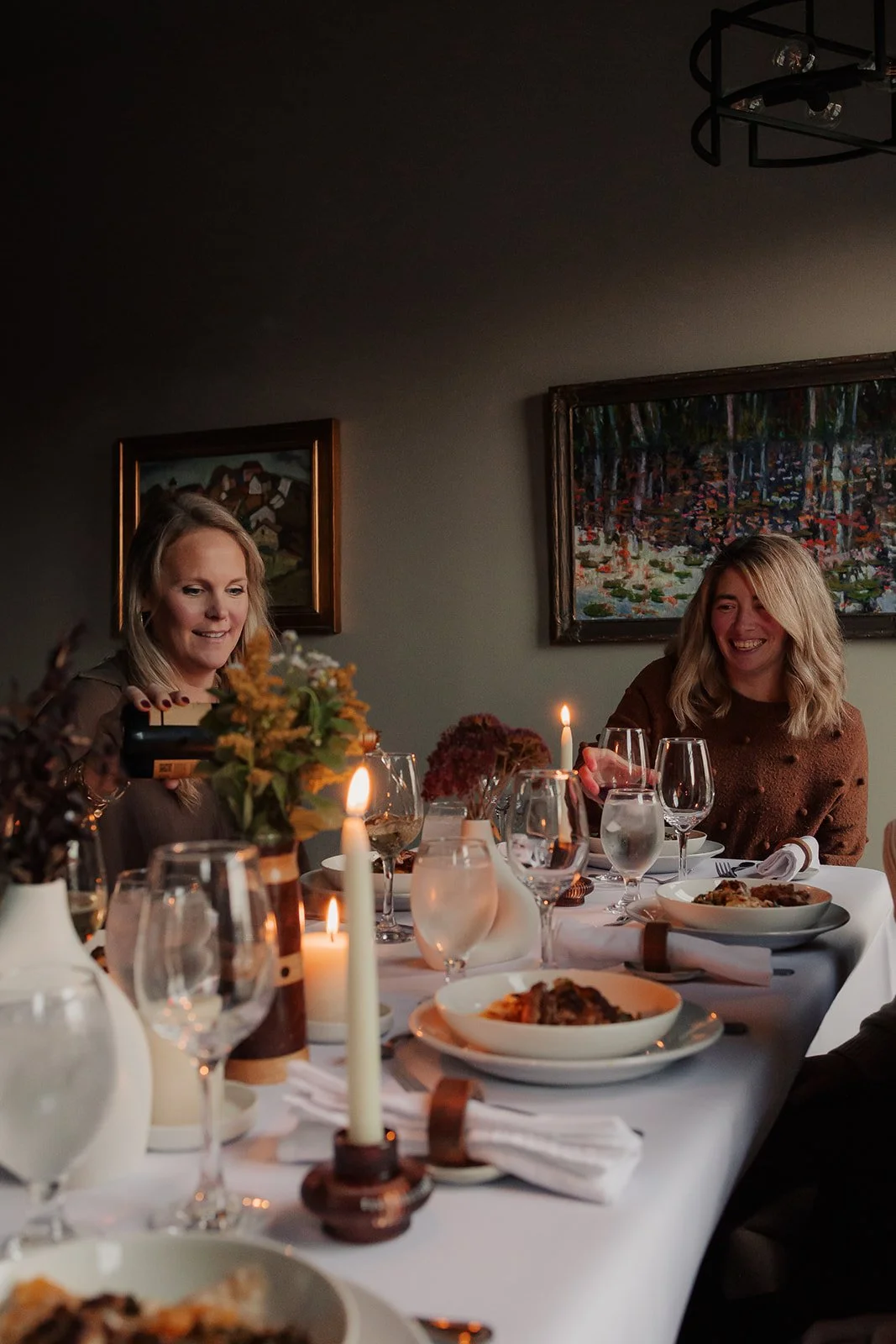 Two women smiling and enjoying dinner at a decorated table with candles and wine glasses in a cozy, art-decorated room.