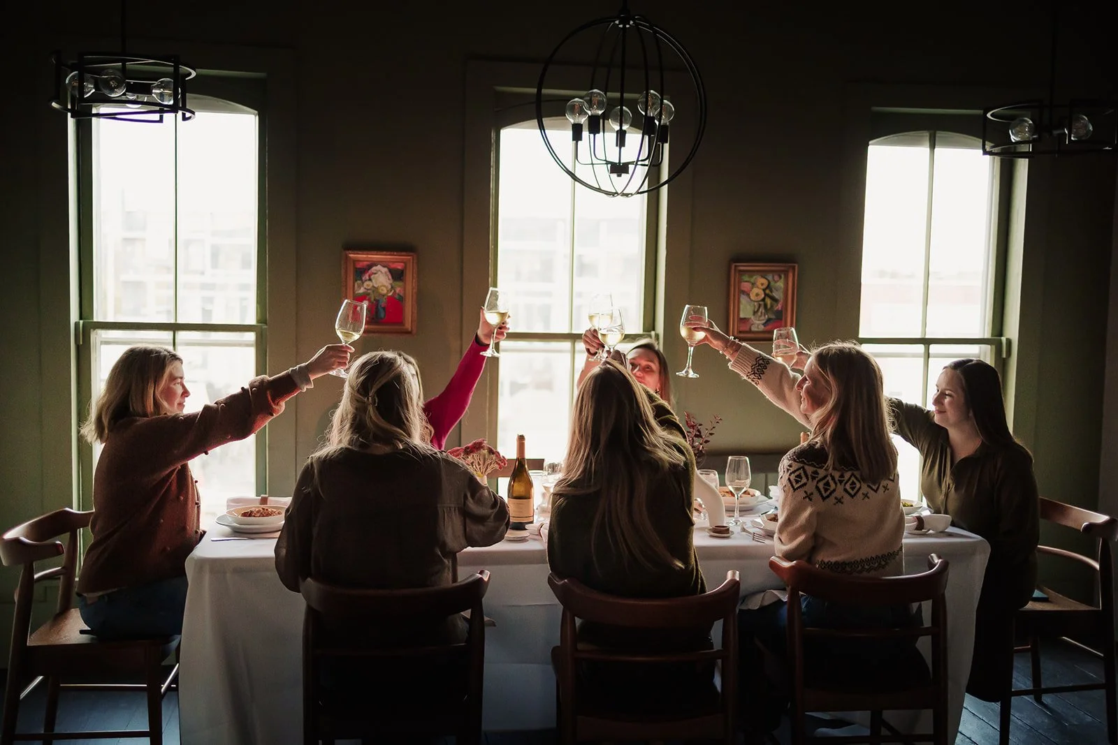 Group of women celebrating and raising glasses of white wine at a dinner gathering in a sunlit room.