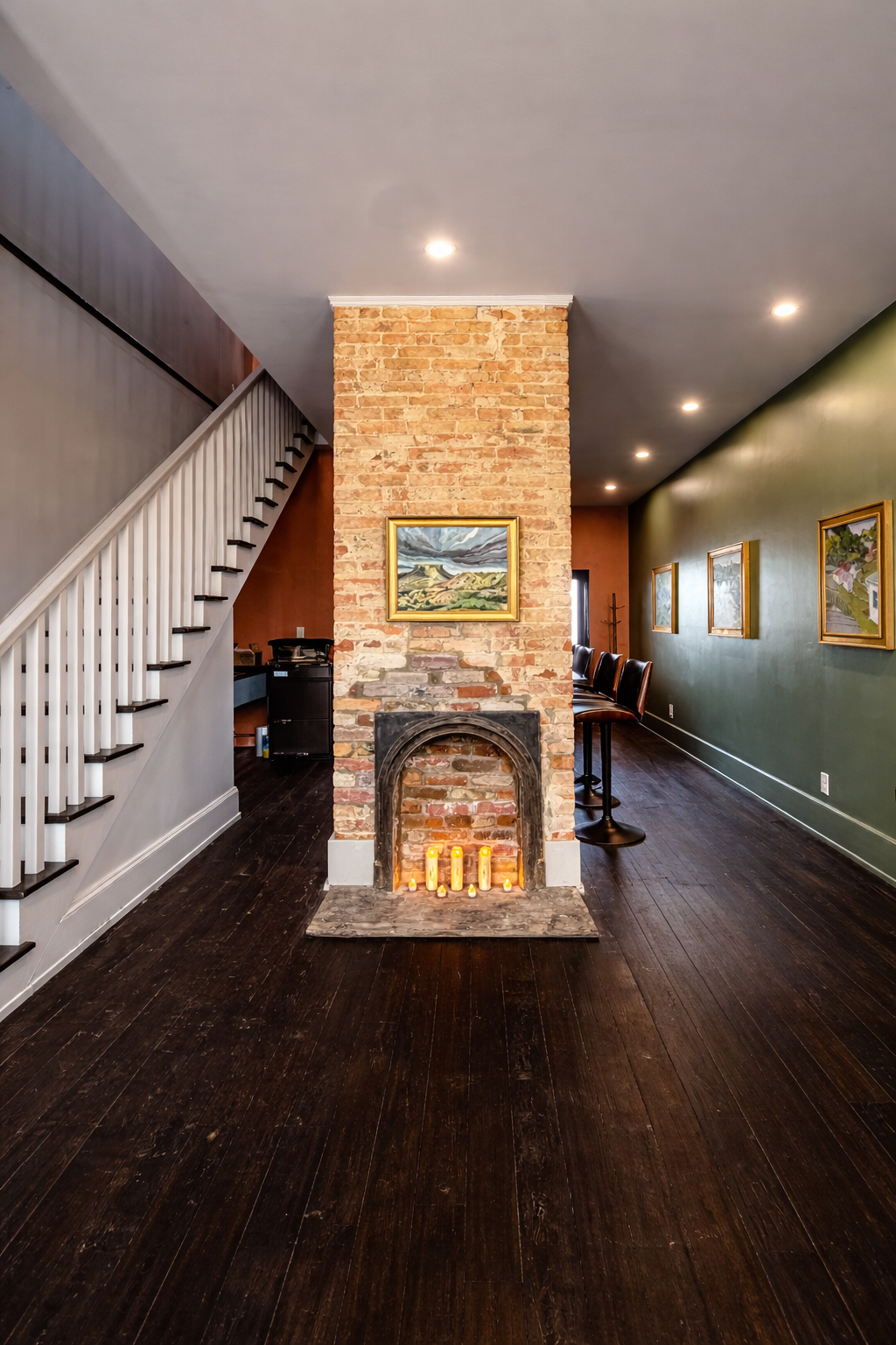 Interior of a living space with a brick fireplace, lit candles inside, dark wooden floor, green wall with framed paintings, staircase with white railing, and bar seating with chairs along the wall.