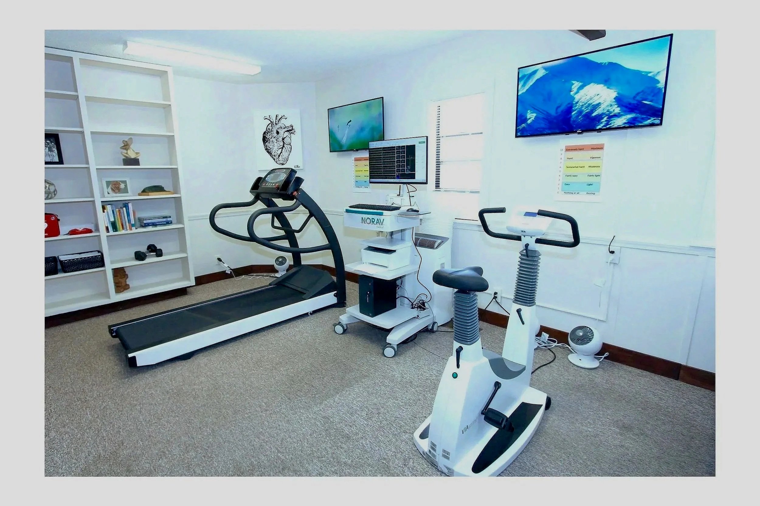Exercise room with treadmill, recumbent bike, and heart rate monitors, with organized shelf and medical equipment, and monitors on the wall.