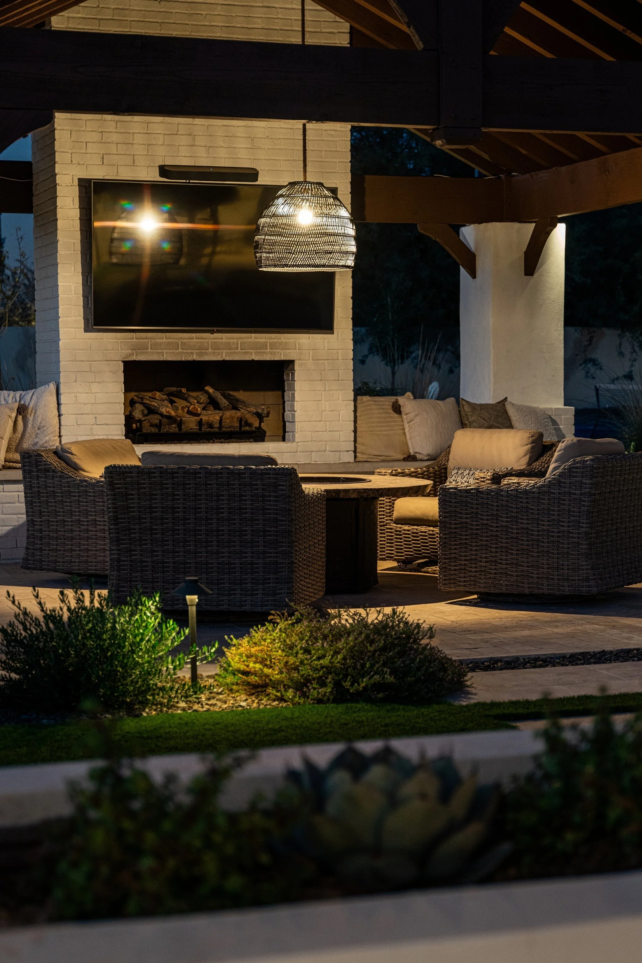Outdoor patio seating area with wicker chairs, a round table, a fireplace, a wall-mounted TV, and ambient lighting at dusk.