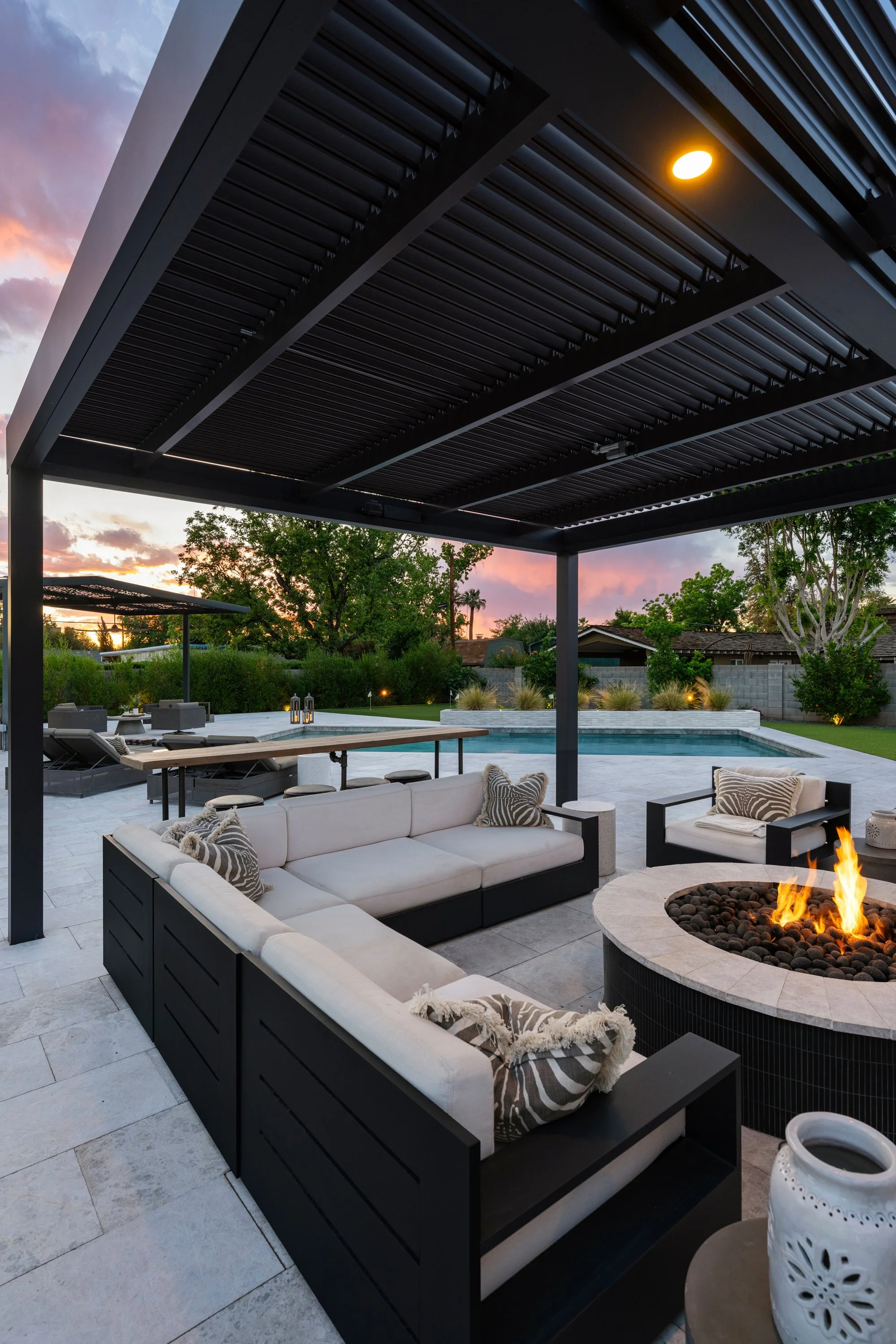 Outdoor patio with modern furniture, a fire pit, swimming pool, and landscaped yard at sunset.