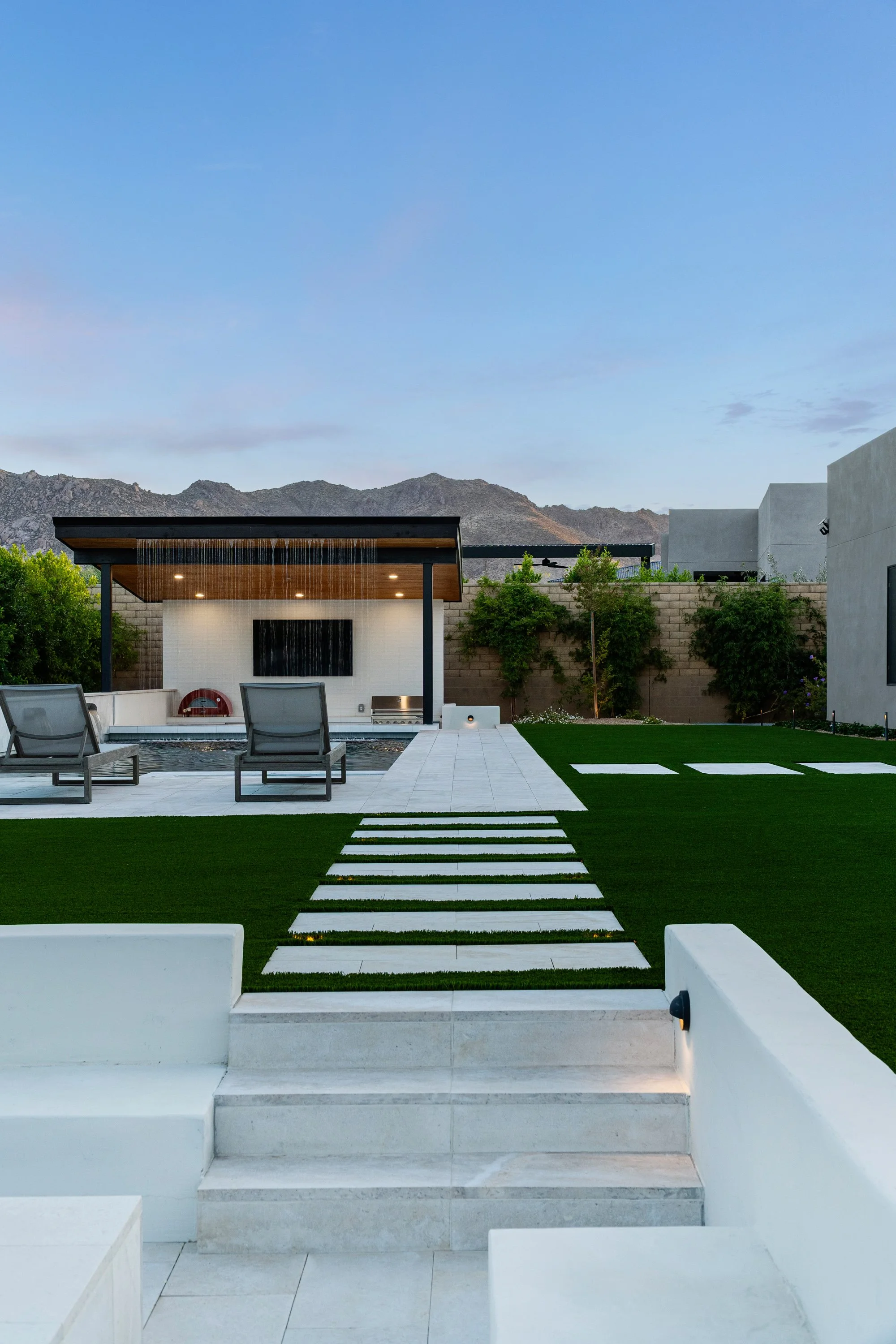 Modern backyard with white steps leading to a green lawn, lounge chairs, a pool, and a shaded outdoor area with a mountain background.