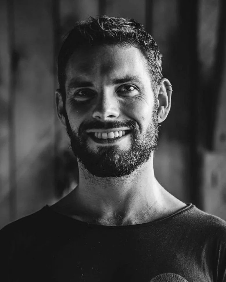 Black and white portrait of a smiling man with a beard, short hair, and an earring on his left ear, wearing a dark shirt.