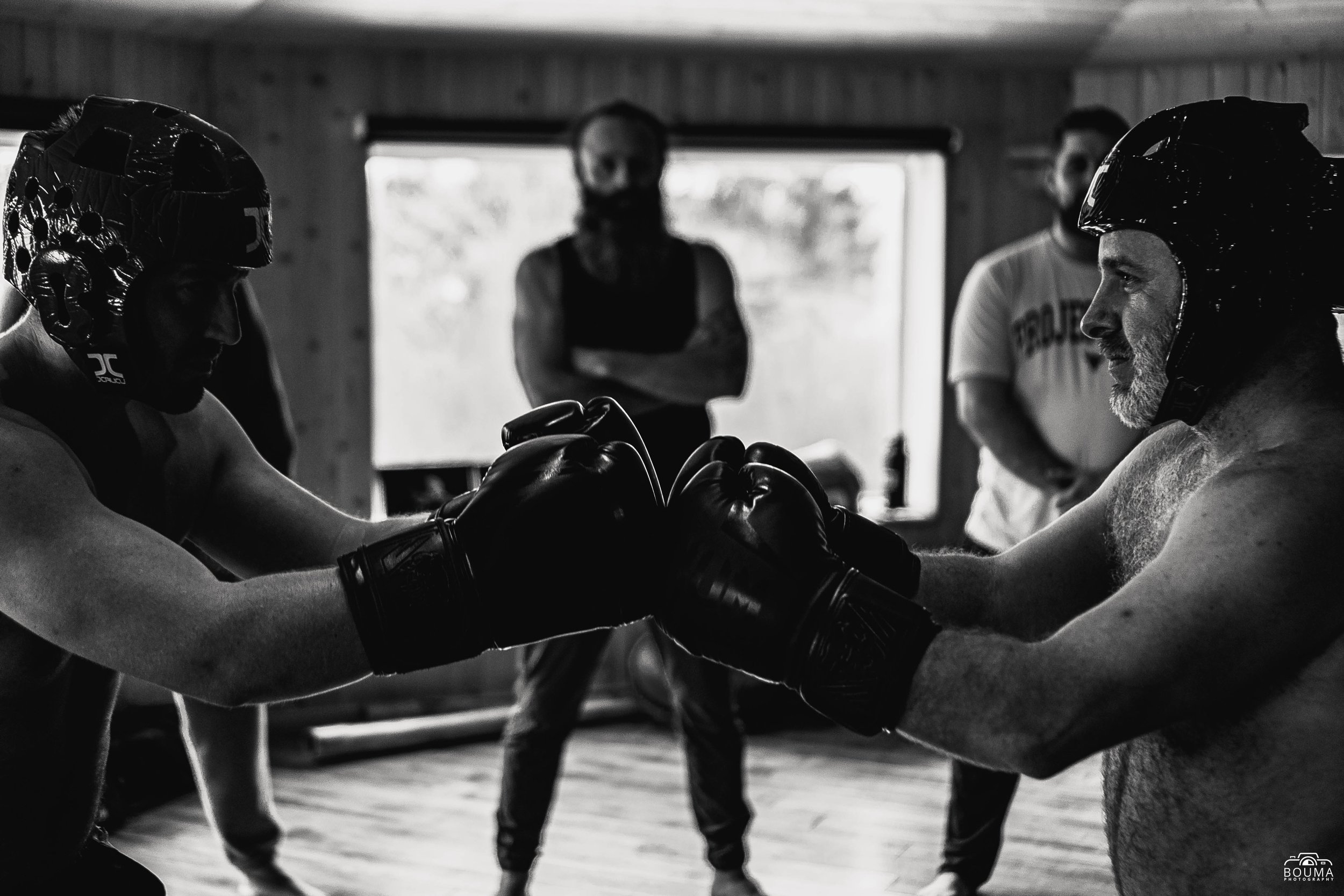 Two shirtless men wearing helmets and gloves are engaging in a fist bump during a boxing training session, with two women observing in the background.