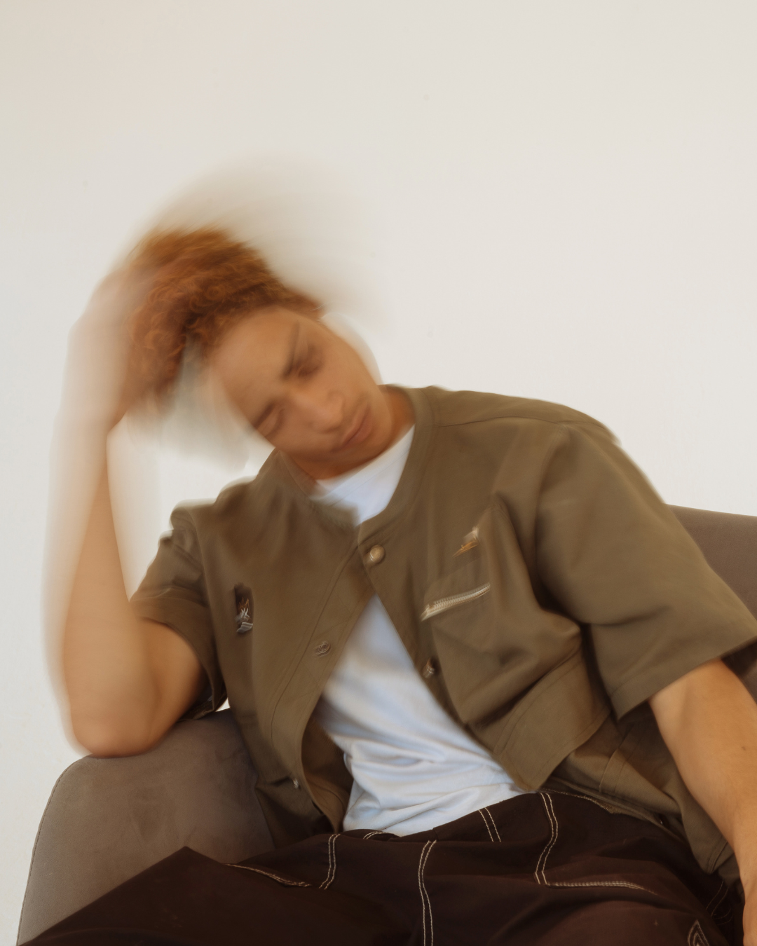 Person with curly hair sitting on a sofa, head tilted, and hand touching their hair, causing motion blur.