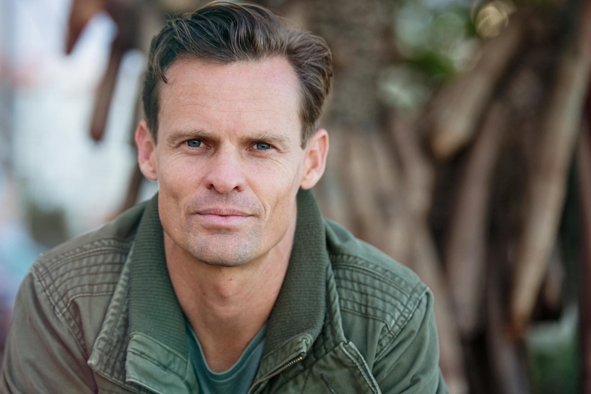A man with short dark hair and blue eyes wearing a green jacket and a blue shirt, outdoors with a defocused background of trees.