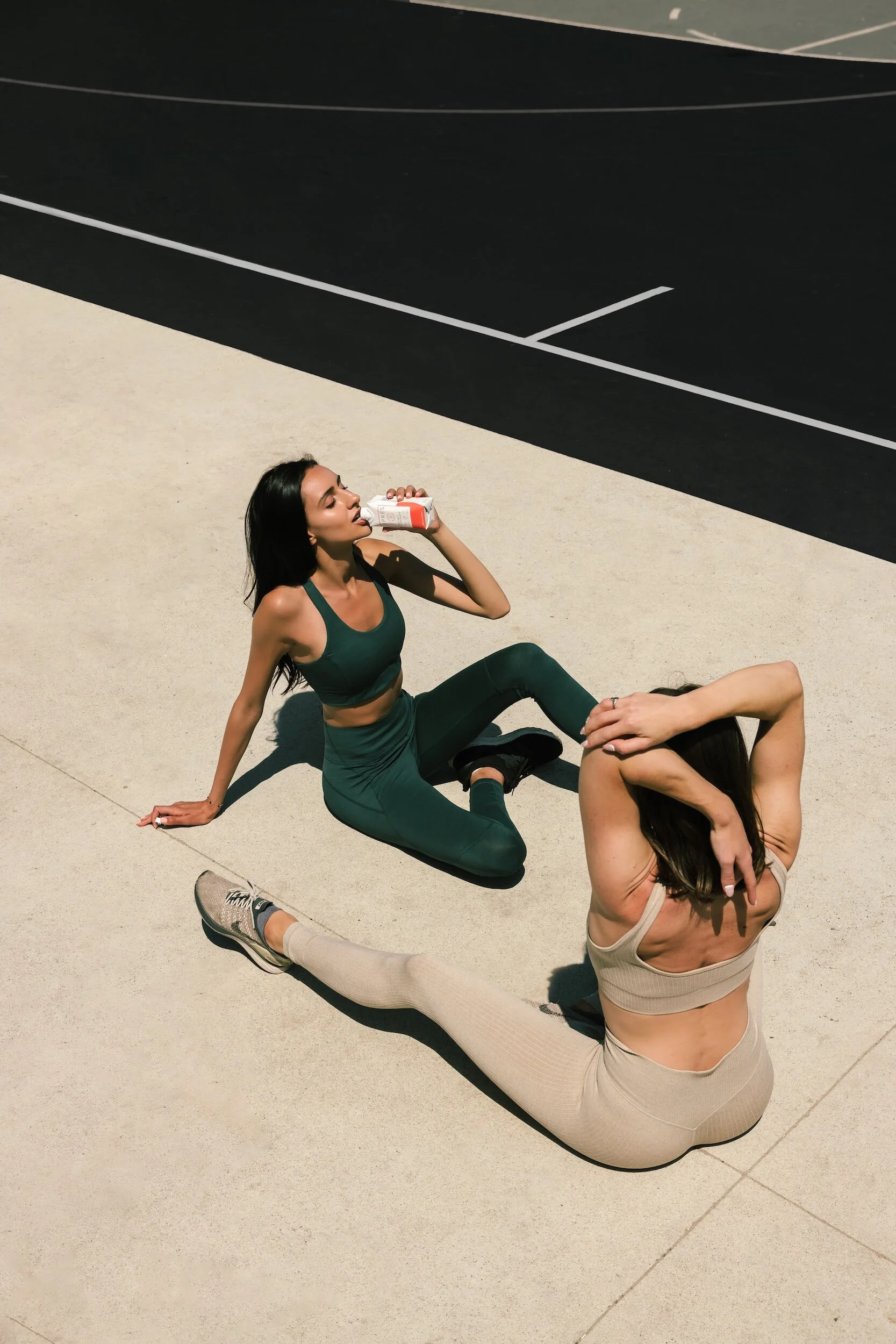 Two women resting on the ground at a sports court, one drinking from a juice box and the other stretching with arms overhead, both in activewear, with a black court and white lines in the background.